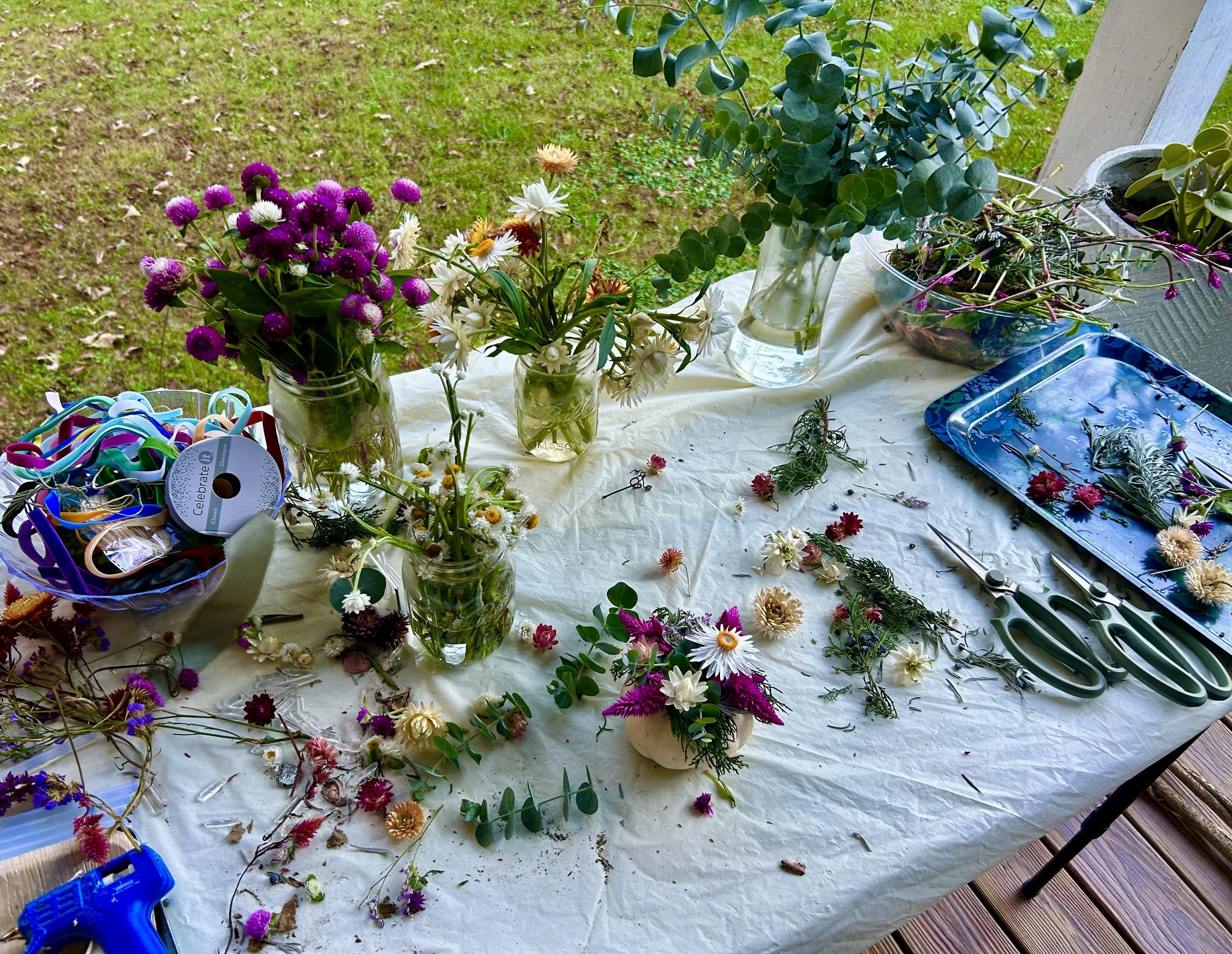 A table covered with various colorful flowers arranged in vases and scattered flower debris, with crafting supplies like scissors, ribbons, and a glue gun nearby. Outside, a grassy yard with there are some potted plants.