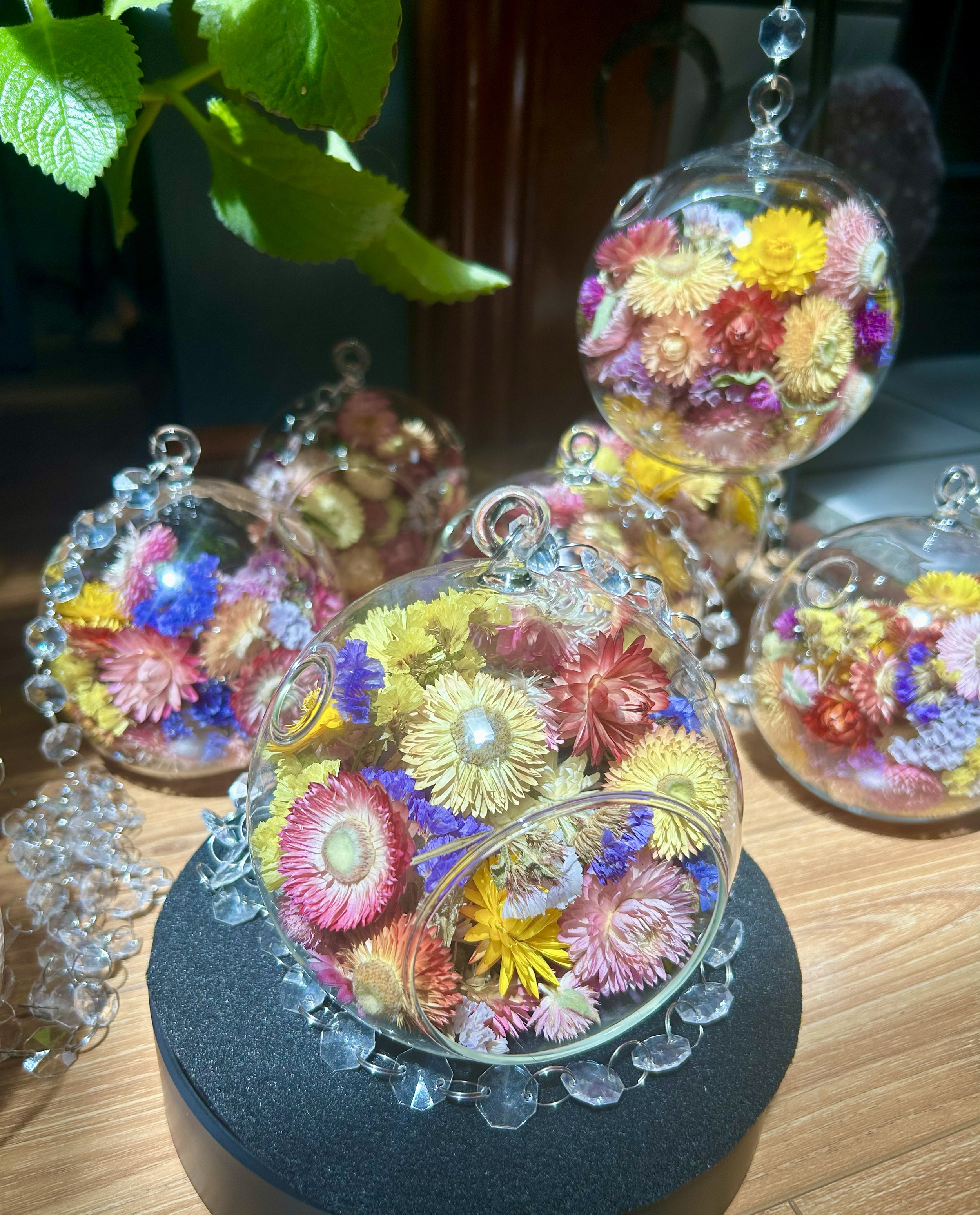 Glass bowl filled with colorful dried flowers including pink, yellow, purple, and cream flowers, and a small portion of green leaves visible on the top left corner. The bowl is placed on a black round stand on a wooden surface.