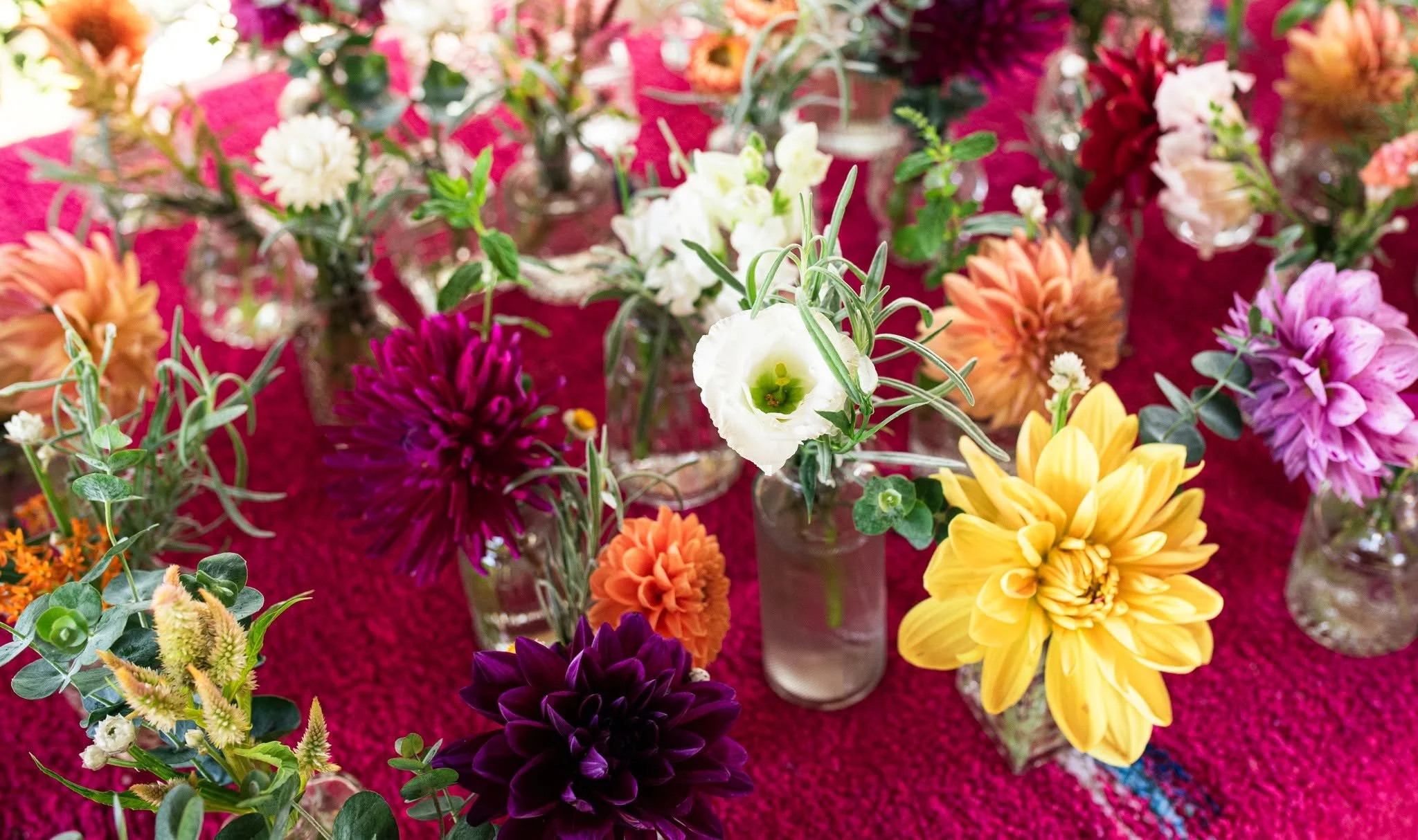 Colorful flower arrangement in glass vases with various flowers including white, purple, yellow, orange, and pink blooms, set on a red textured surface.