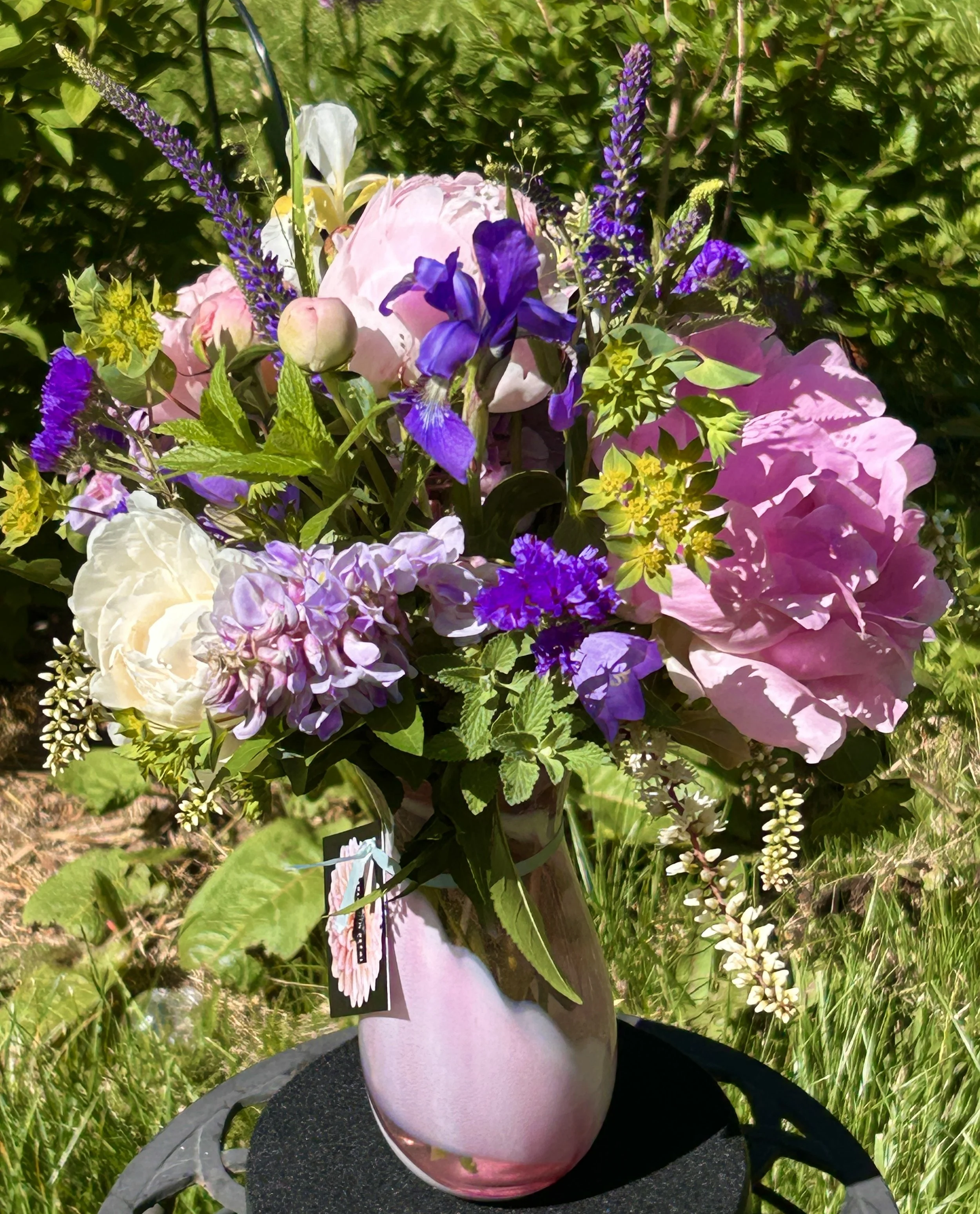 Vase with pink roses, purple irises, and other flowers on a black table outside with greenery in the background.
