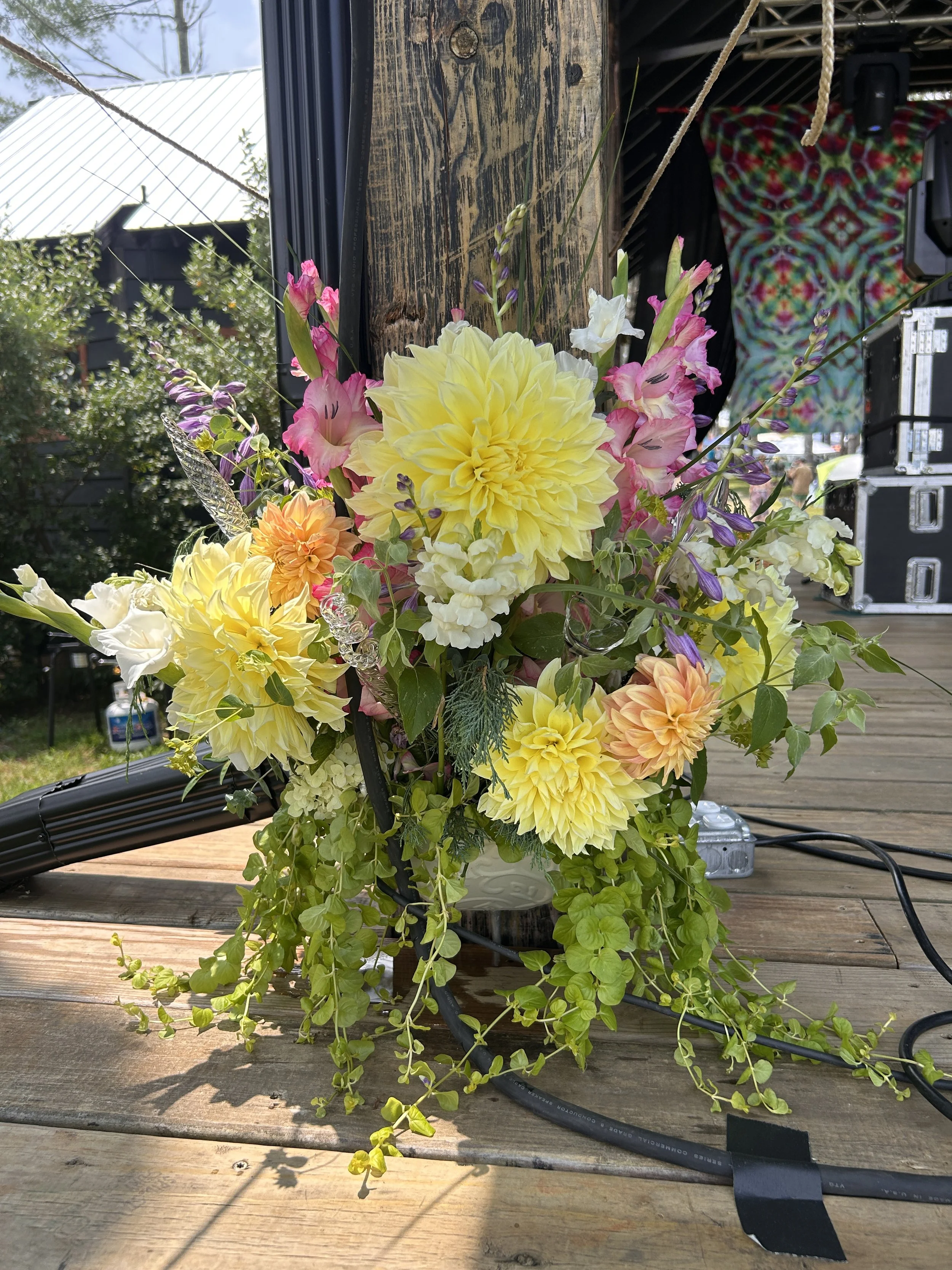 A colorful bouquet of flowers including yellow dahlias, pink gladiolus, purple statice, and trailing green vines, arranged on a wooden surface with a rustic wooden post and a background of outdoor structures and greenery.