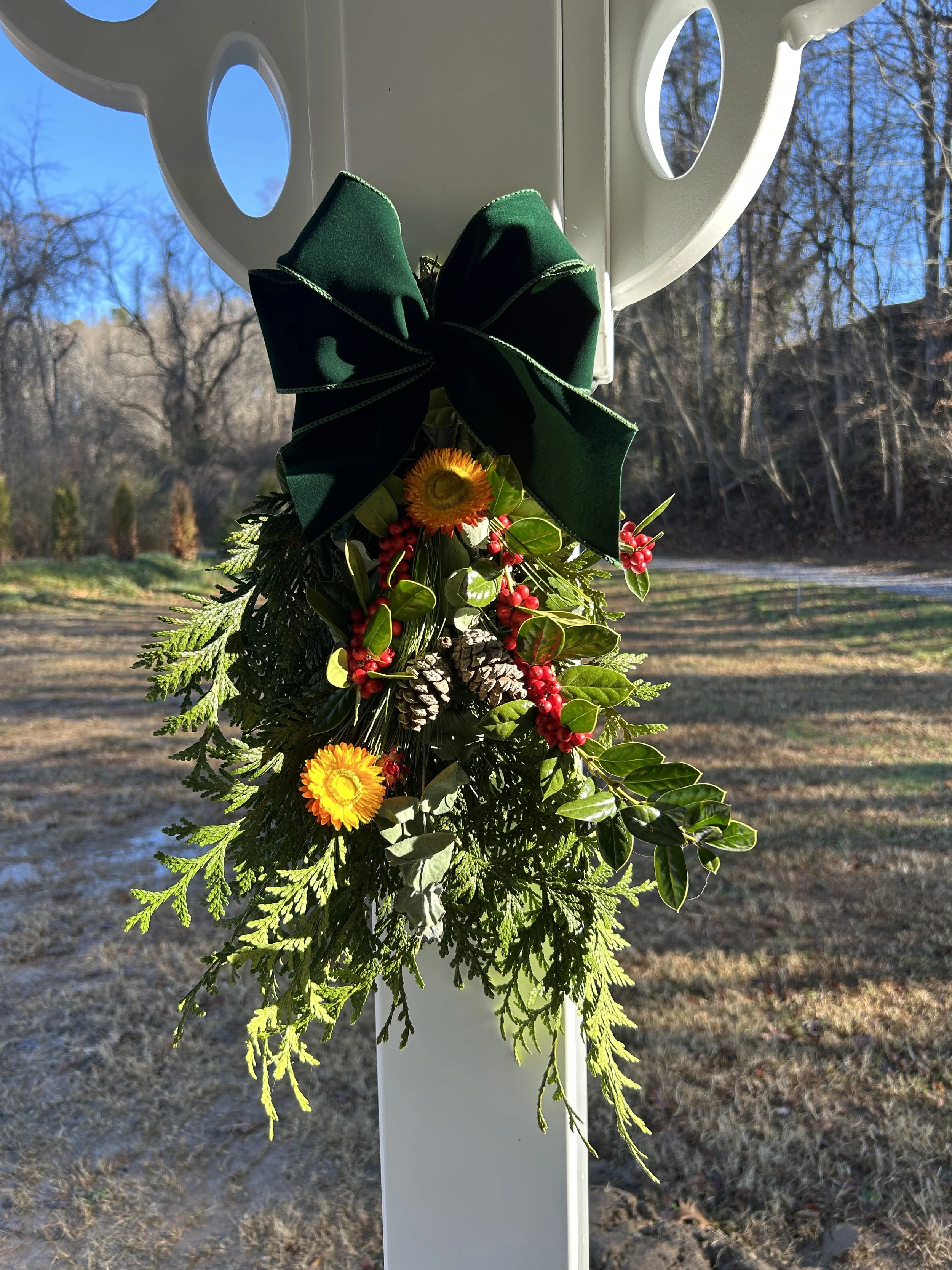 A holiday decoration with a large green bow, yellow sunflower, red berries, pinecones, and various greenery attached to a white post outdoors during daytime.