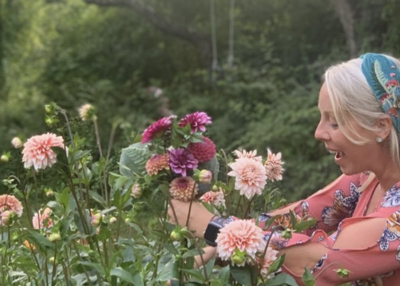 A woman with blonde hair and a colorful headscarf smiling while picking pink and purple flowers in a garden.