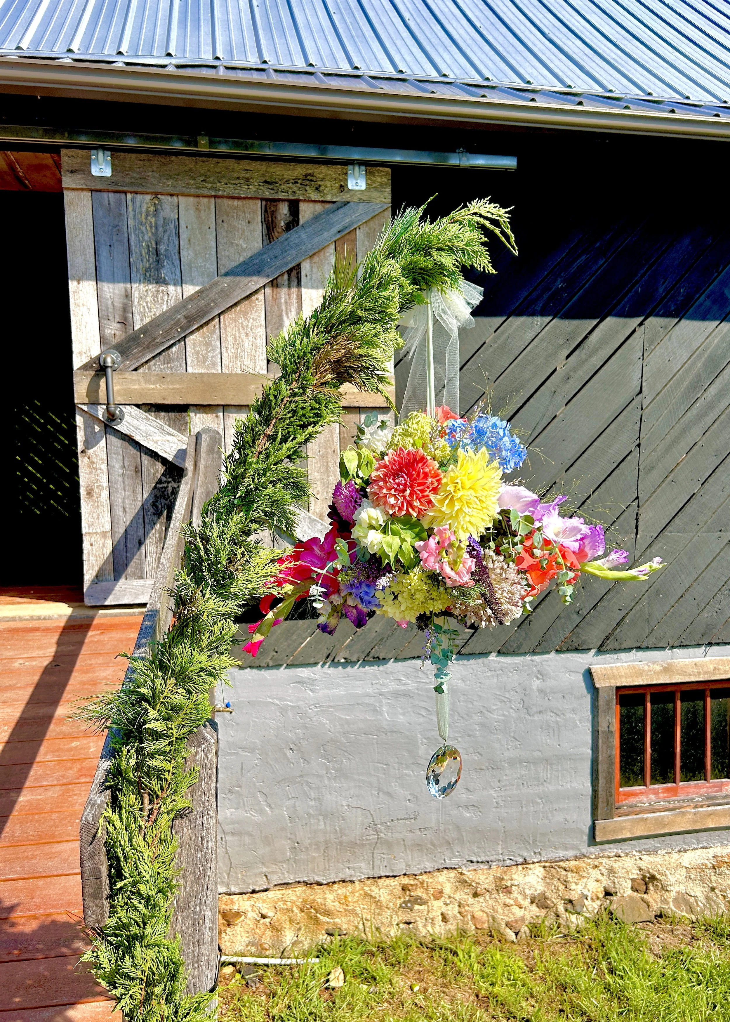 A decorative garden arrangement against a rustic building wall with a wooden door. It features green foliage, colorful flowers, and a hanging crystal ornament