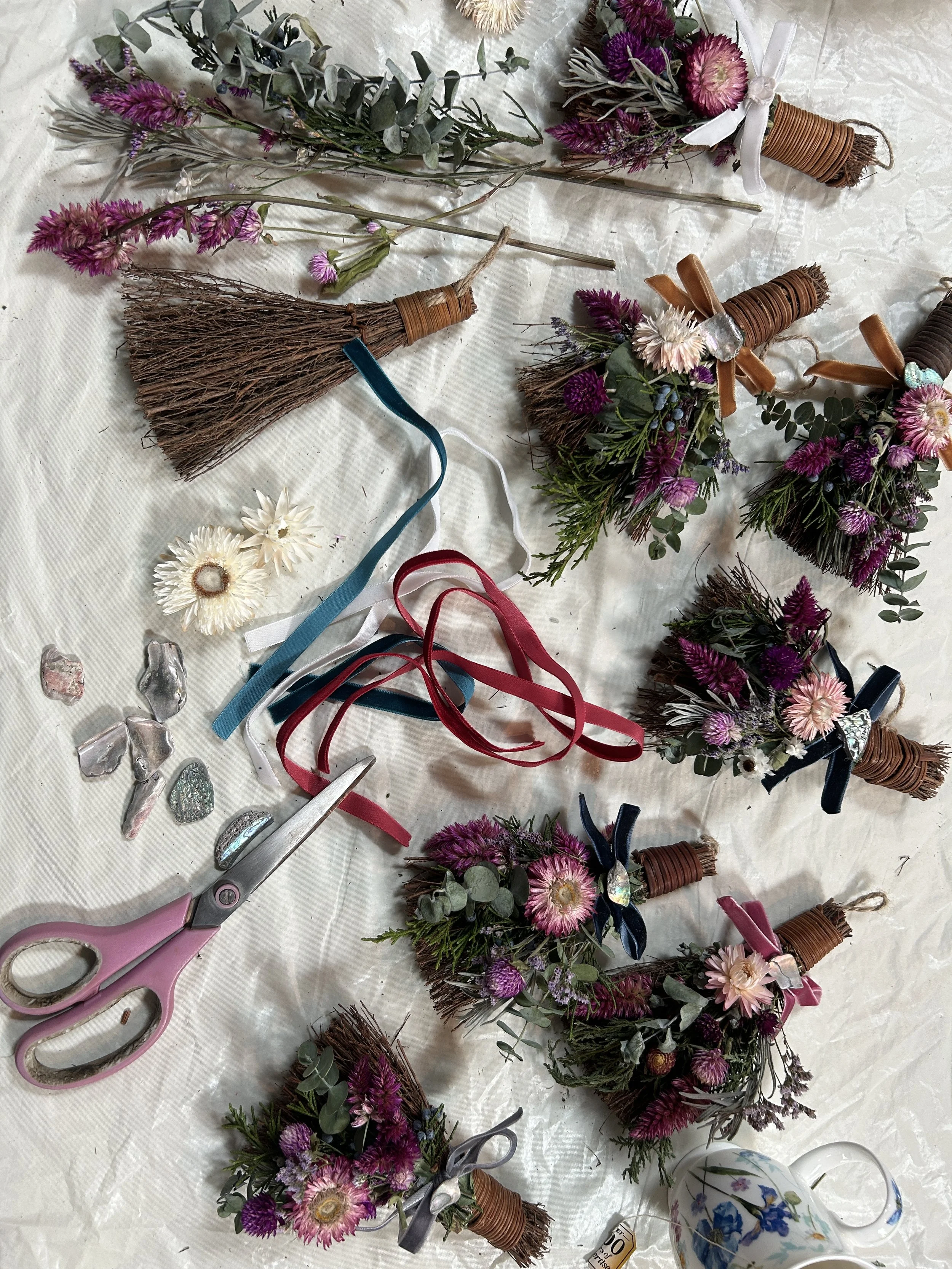 Various artificial flower bouquets with pink, purple, and white flowers, some with ribbons and wire in a craft workspace, along with a pair of pink scissors and a porcelain mug with blue flowers.