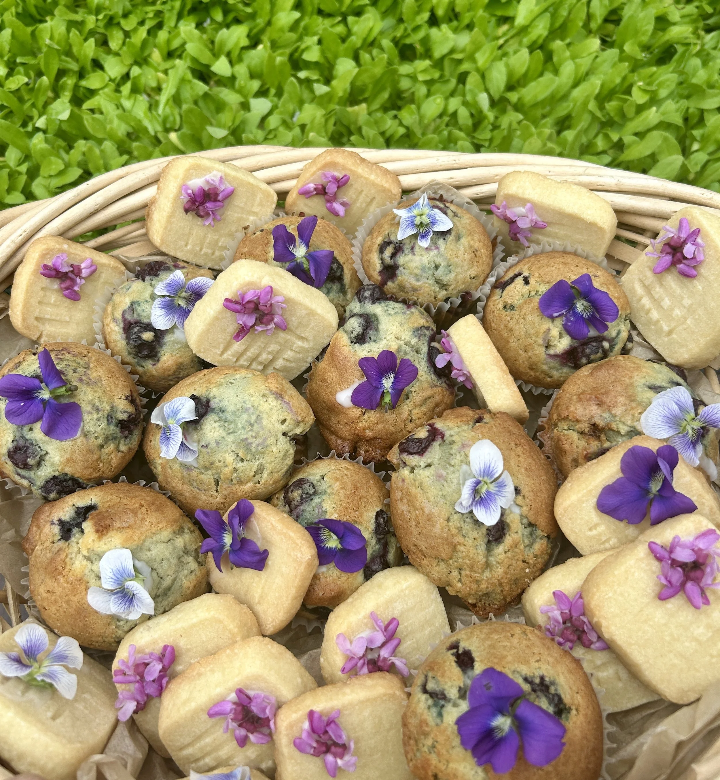 Assorted muffins decorated with edible flowers in a wicker basket, set against a background of green leaves.