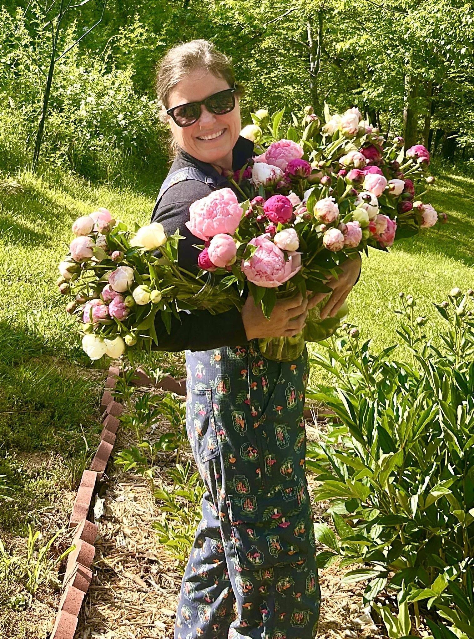 A woman smiling and holding a large bouquet of pink and white peonies outdoors on a sunny day, with lush green trees in the background.