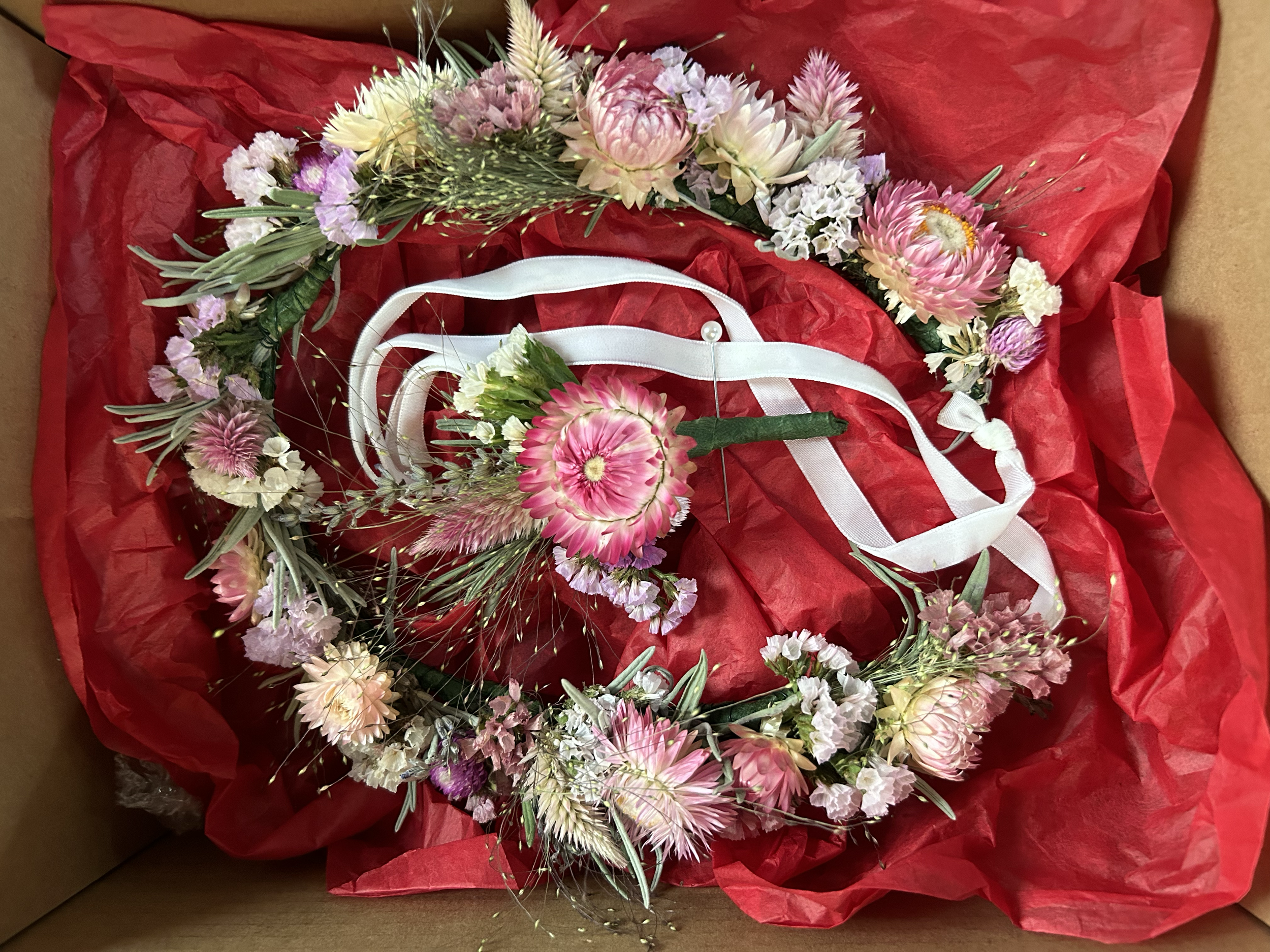 A floral crown made of pink, white, and purple flowers resting on red tissue paper in a cardboard box.