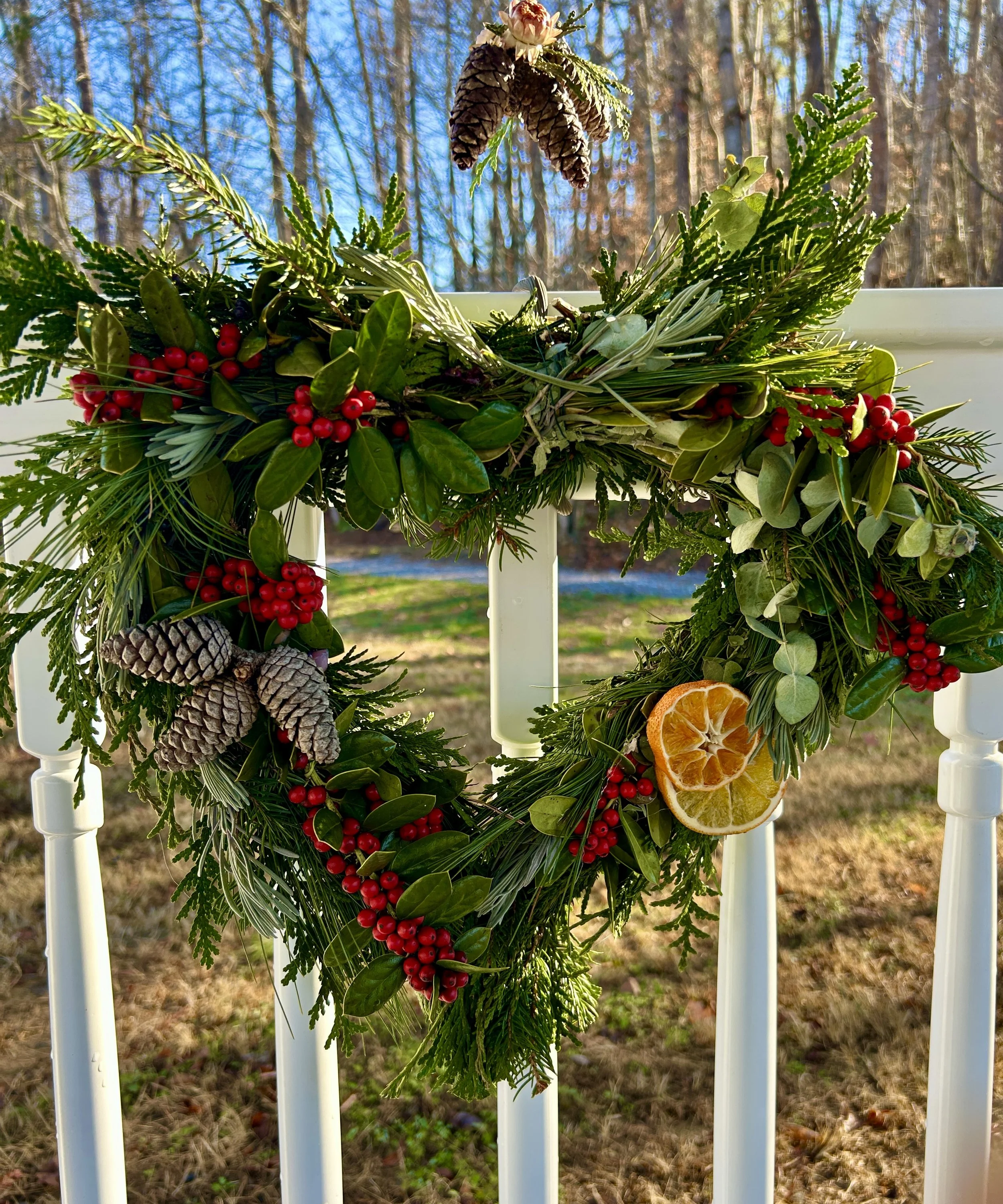 A Christmas wreath decorated with pinecones, red berries, green leaves, and slices of dried orange, hanging on a white porch railing outdoors on a sunny winter day.