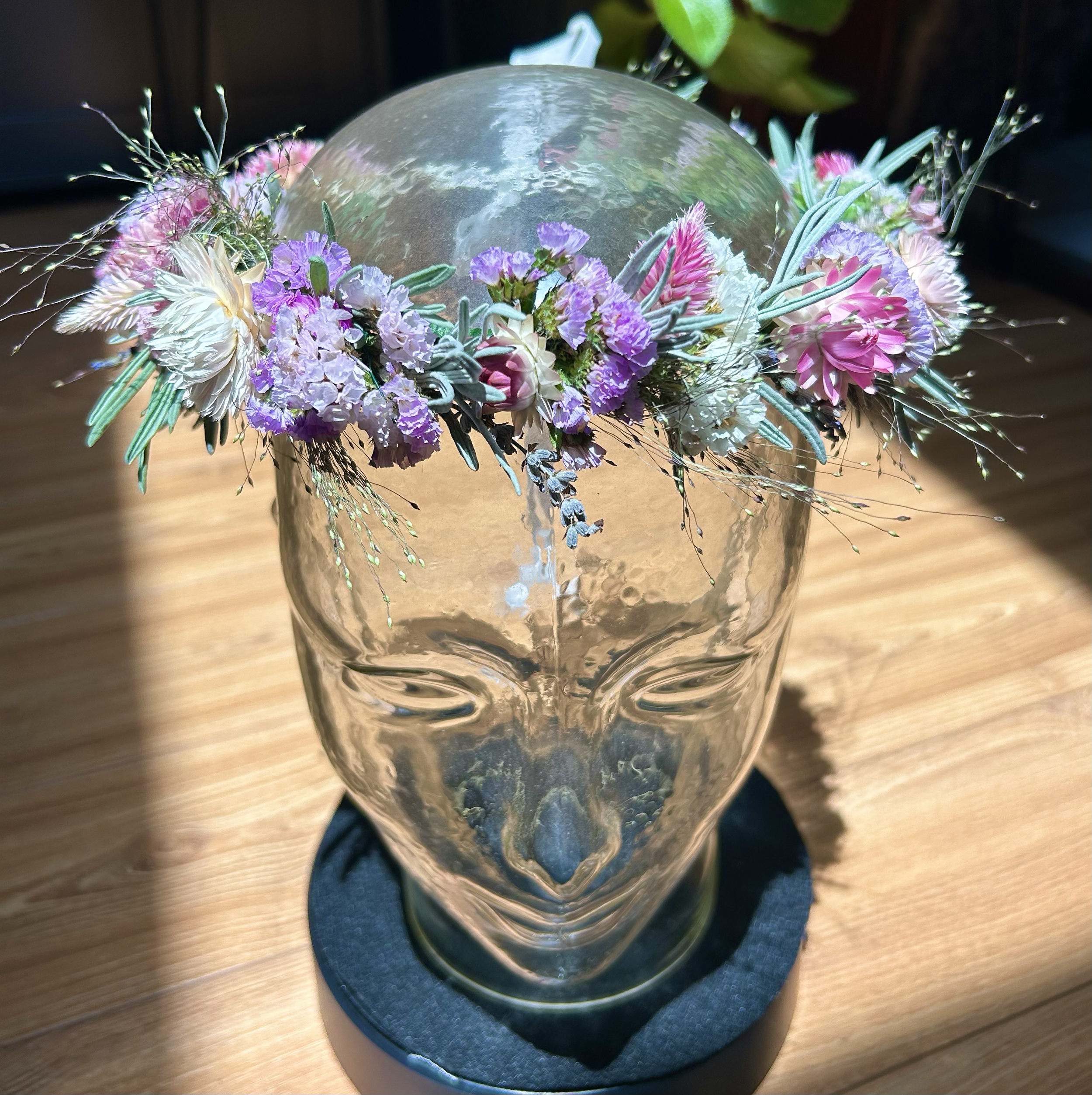 A glass vase with a round shape and textured surface holds a floral arrangement of purple, pink, white, and green flowers and foliage. The vase is placed on a piece of dark fabric on a wooden surface.
