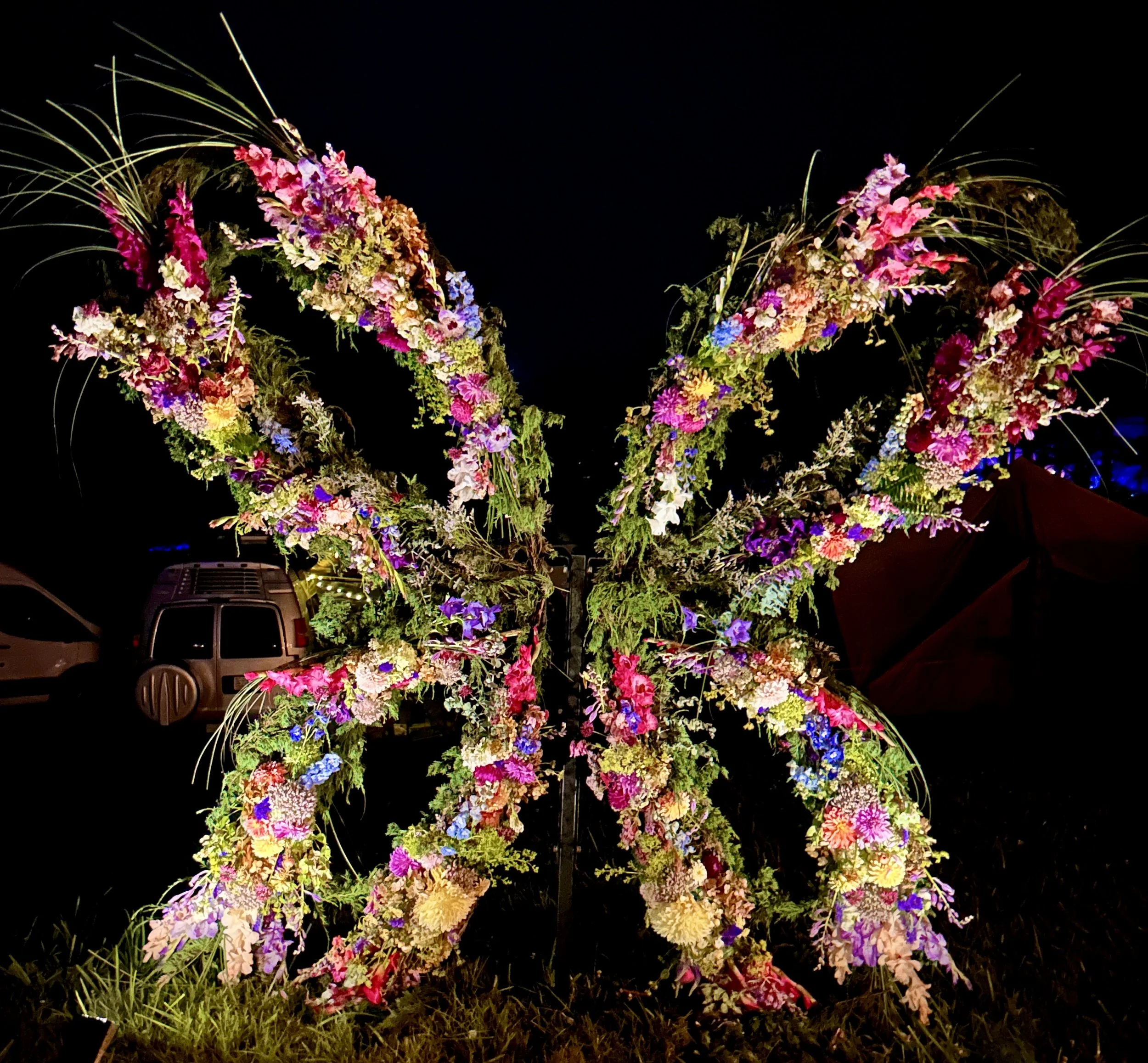 Colorful flower arrangement illuminated at night, with various pink, purple, white, and yellow blooms, and green foliage, set against a dark background.