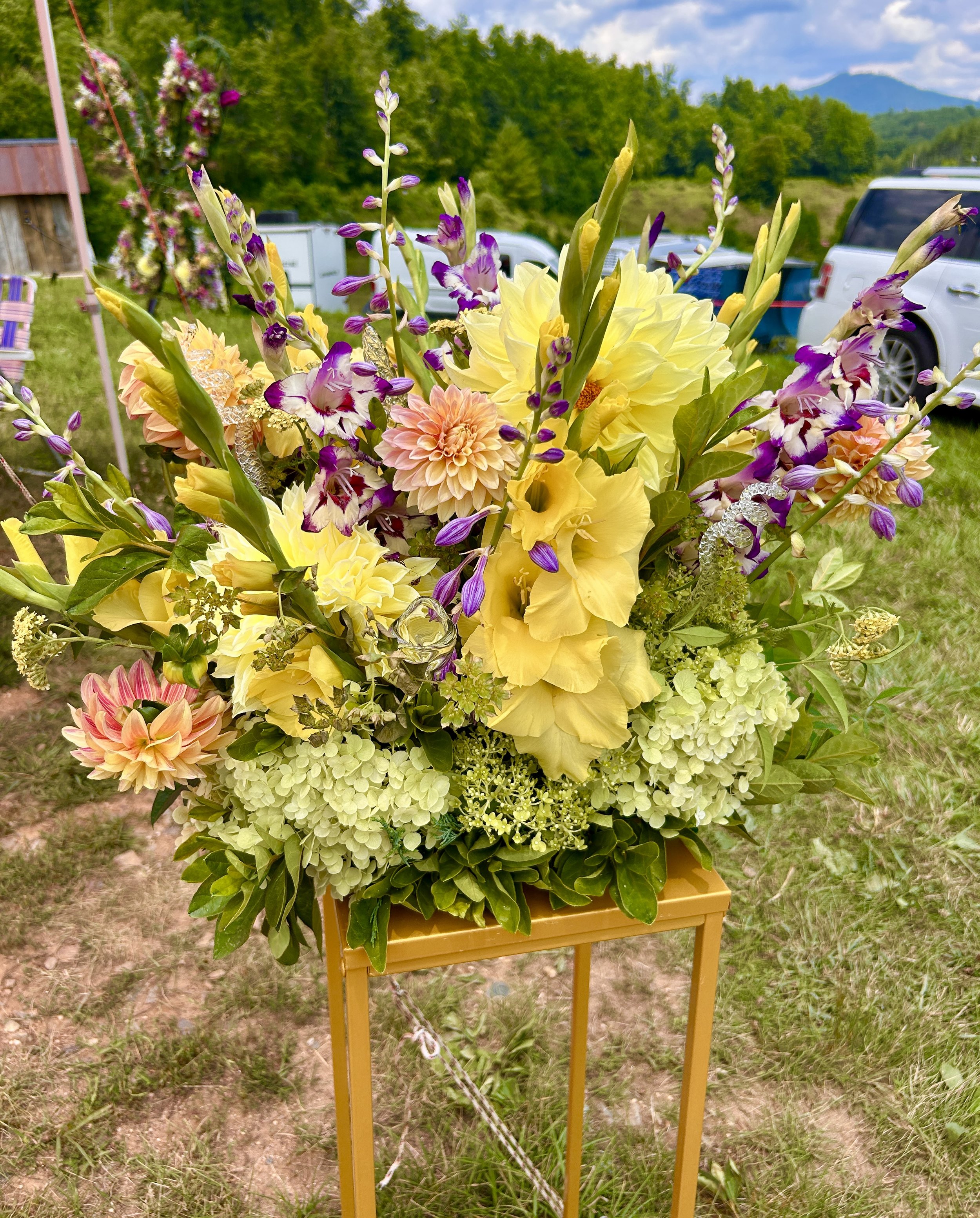 A large bouquet of mixed flowers, including yellow, pink, purple, and white blooms, arranged on a yellow stand outside on grass with cars and green trees in the background under a partly cloudy sky.