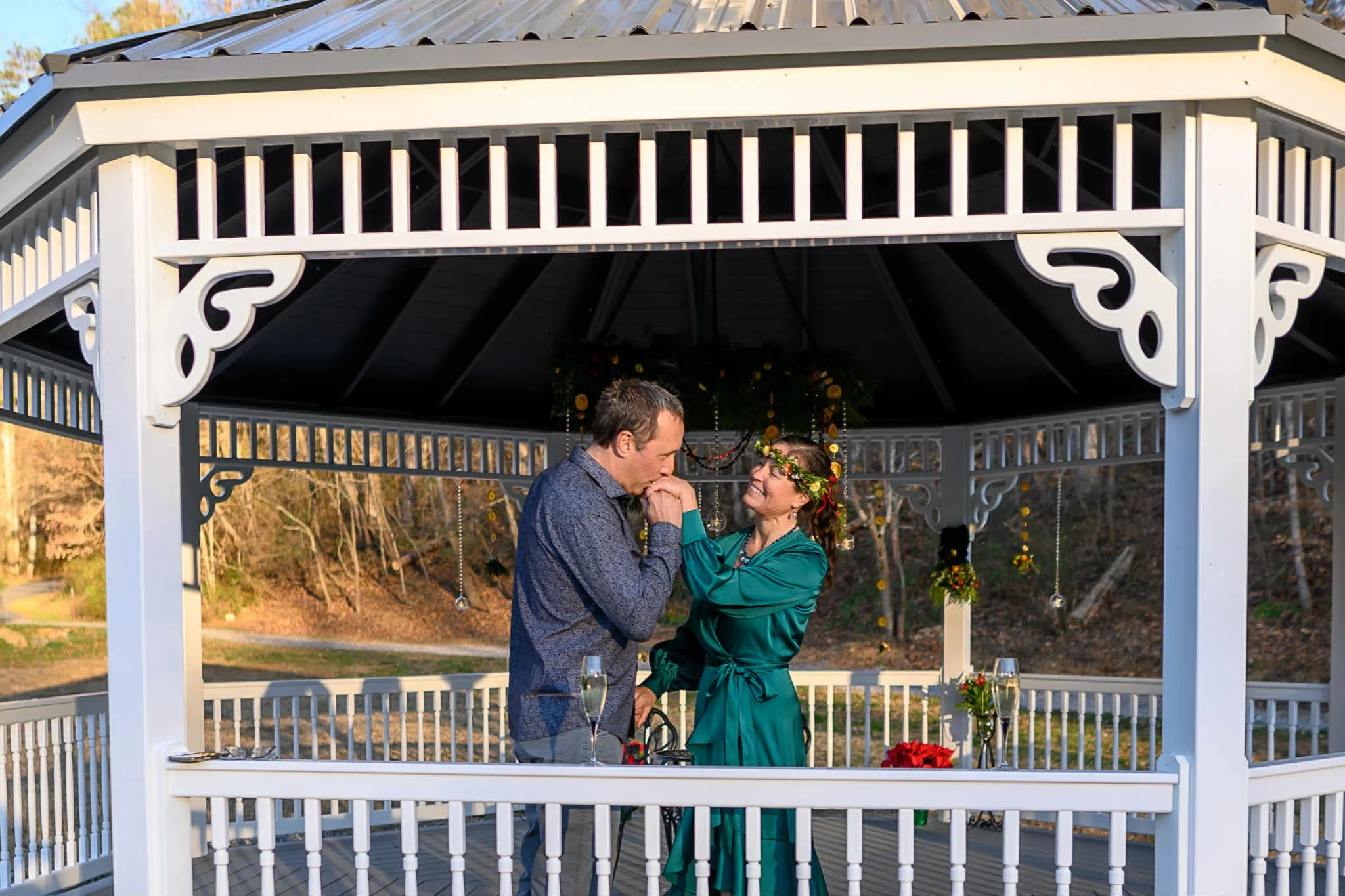 A couple is exchanging rings in a gazebo, celebrating a wedding ceremony outdoors, surrounded by trees and decorated with flowers.