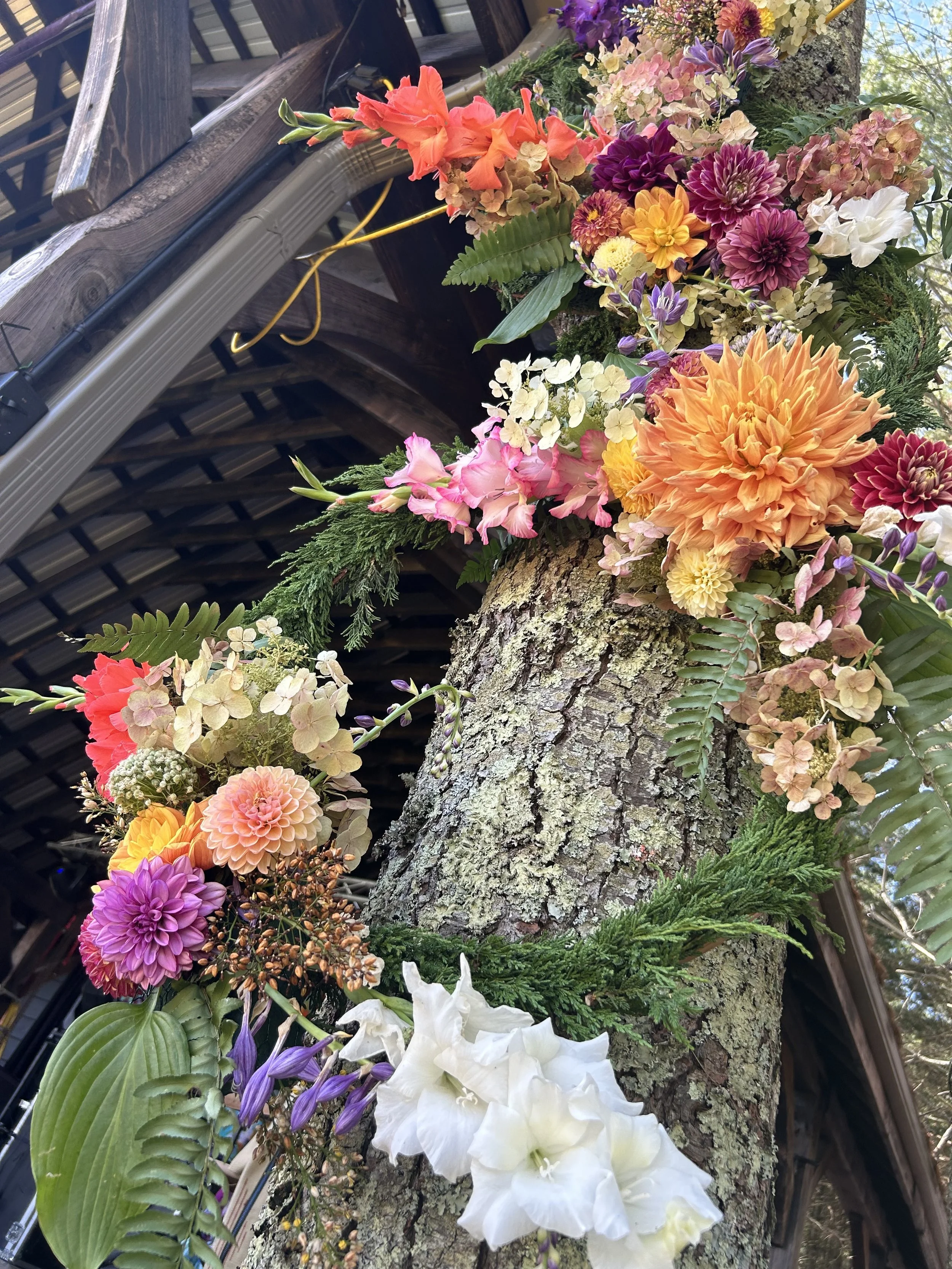 A tree trunk with a variety of colorful flowers and lush green foliage arranged around it, all under a wooden structure with a partially visible corrugated metal roof.