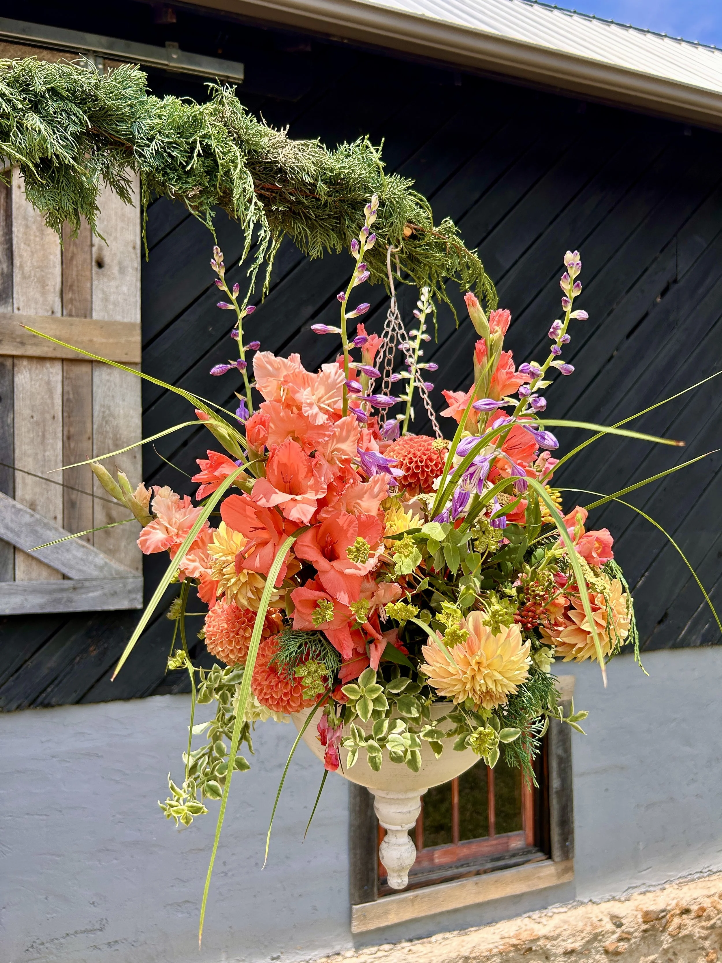 A decorative flower arrangement in a white pedestal vase placed outside on a white wall near a window and black fence, featuring pink, orange, and yellow blooms with green foliage and purple accents.
