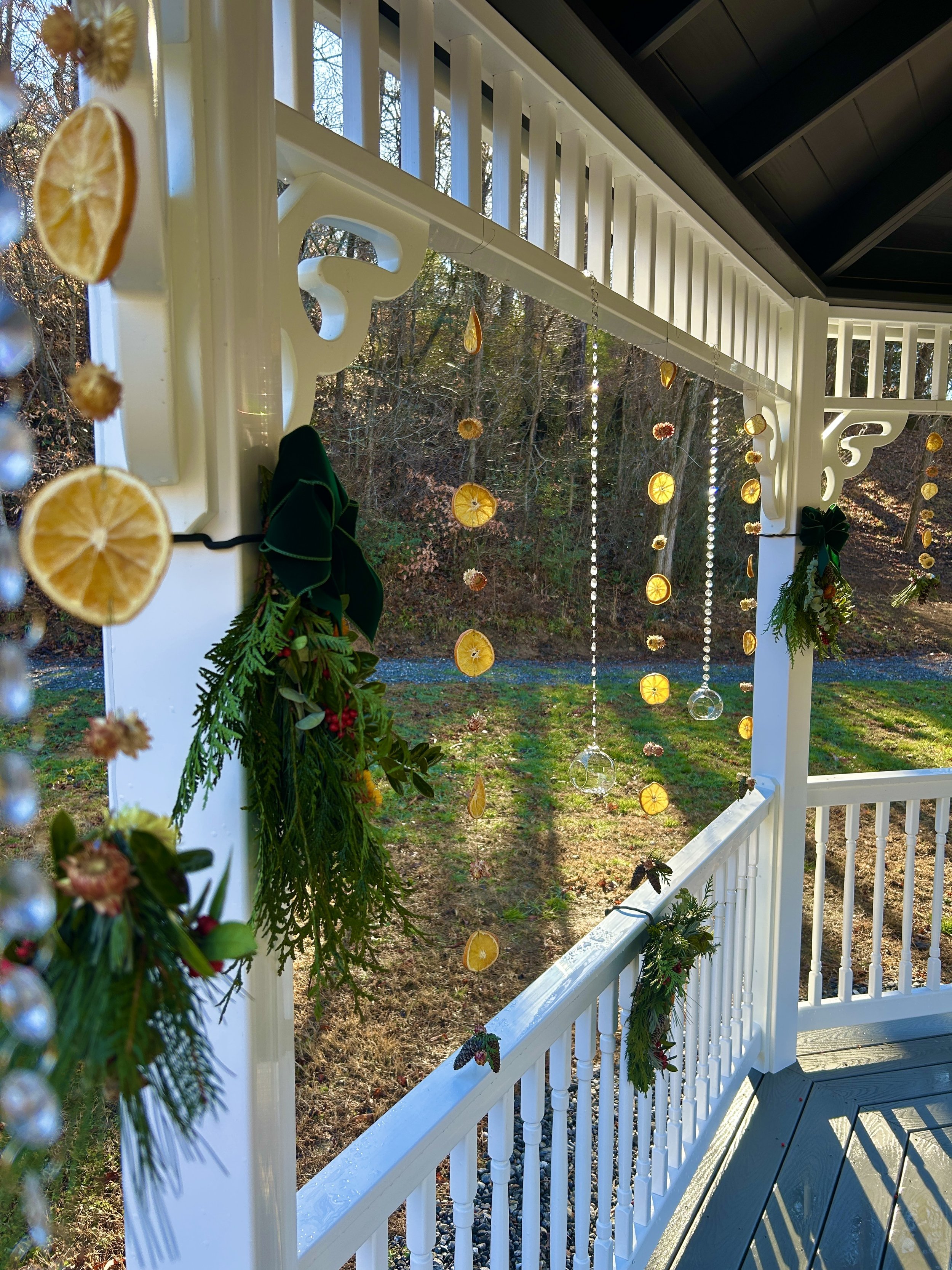 A decorated porch with hanging dried orange slices and small flowers, with white lattice and railing, overlooking a backyard with trees and grass in the background.