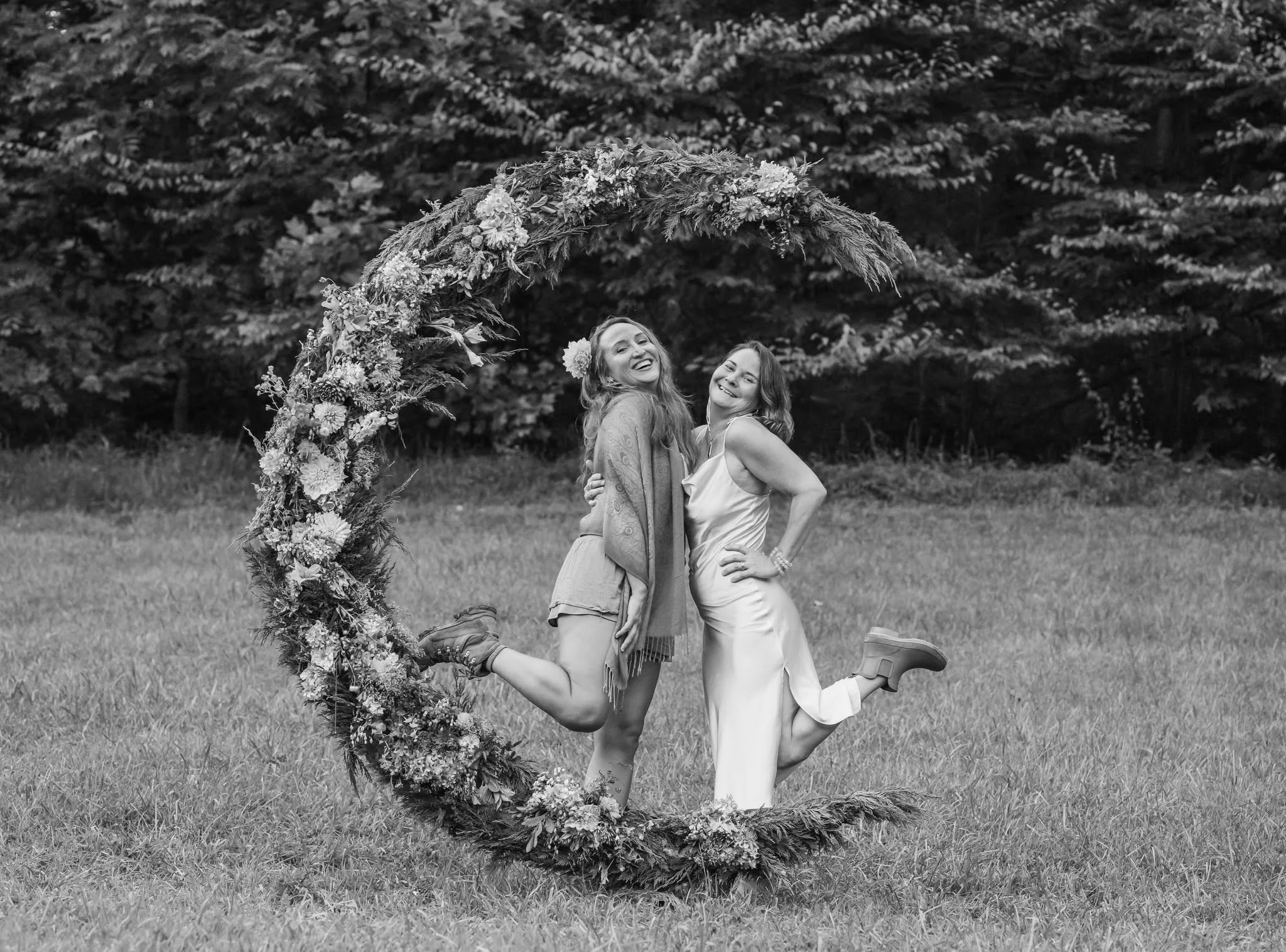 Two women smiling and embracing each other, one with a flower in her hair, standing in front of a floral arch outdoors in a grassy area.