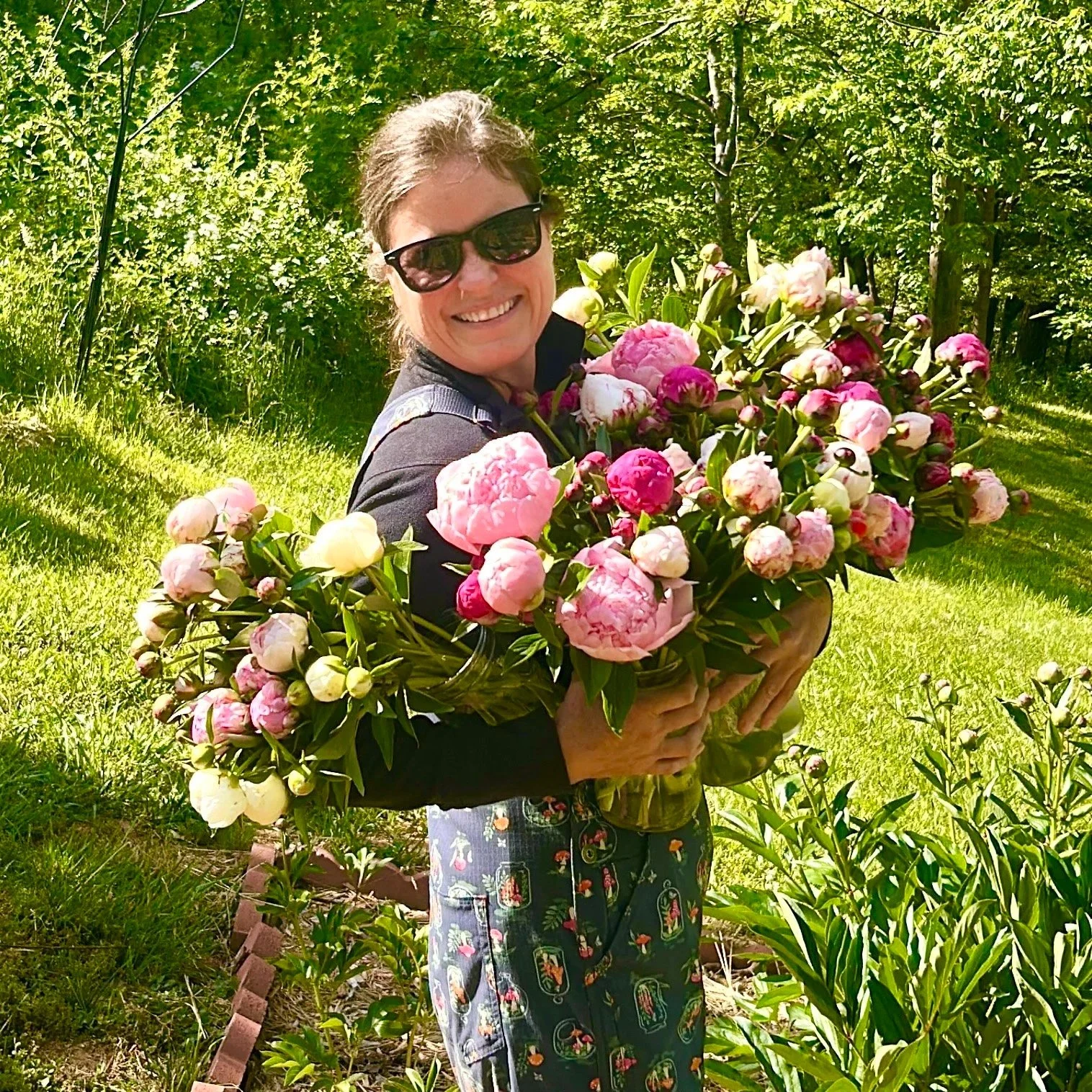 Woman holding a large bouquet of pink and white peonies in a garden with green trees and grass.