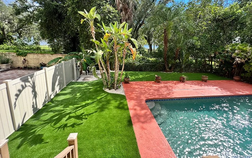 A backyard featuring a swimming pool, green artificial grass, tropical plants, and potted plants on a red concrete patio.