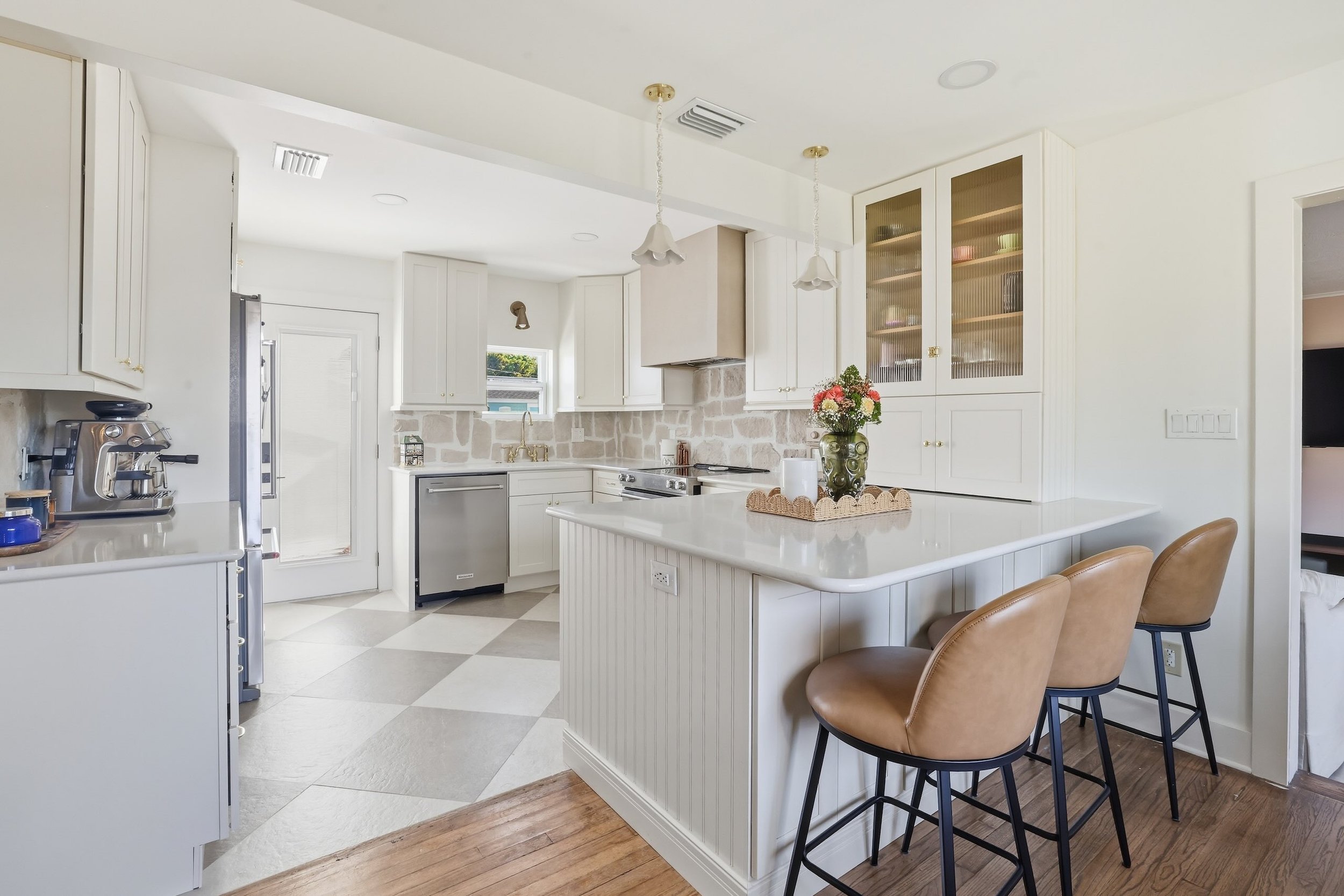 A white kitchen with a stone backsplash, white cabinets, a modern faucet, and a white island countertop. There are two brown bar stools at the island, a vase with flowers, and a few kitchen appliances.