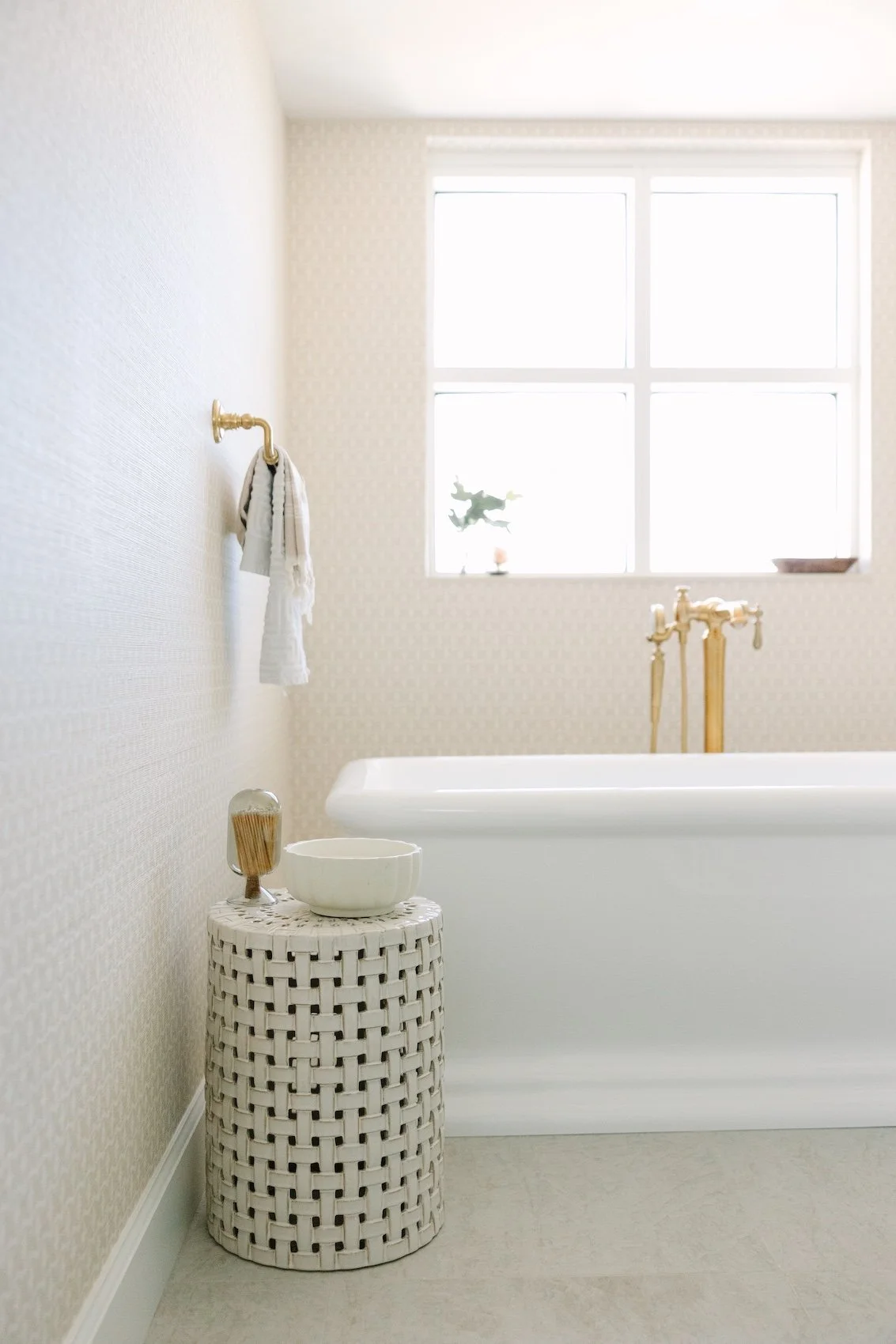 A bright and airy bathroom with a window, white bathtub, gold fixtures, a woven cylindrical side table with a bowl and a jar with toothbrushes, and a towel hanging on a wall hook.