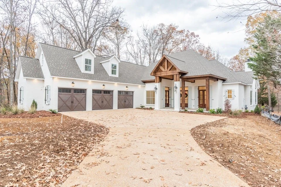 New modern house with a three-car garage, white exterior walls, and a large wooden porch entrance. The driveway is unpaved and surrounded by fallen leaves, with trees and overcast sky in the background.