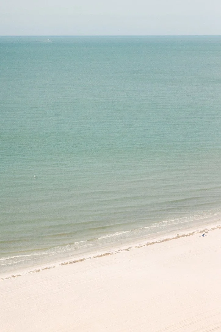A peaceful beach scene with turquoise waters and white sandy beach, minimal waves, and a person sitting alone on the shore.