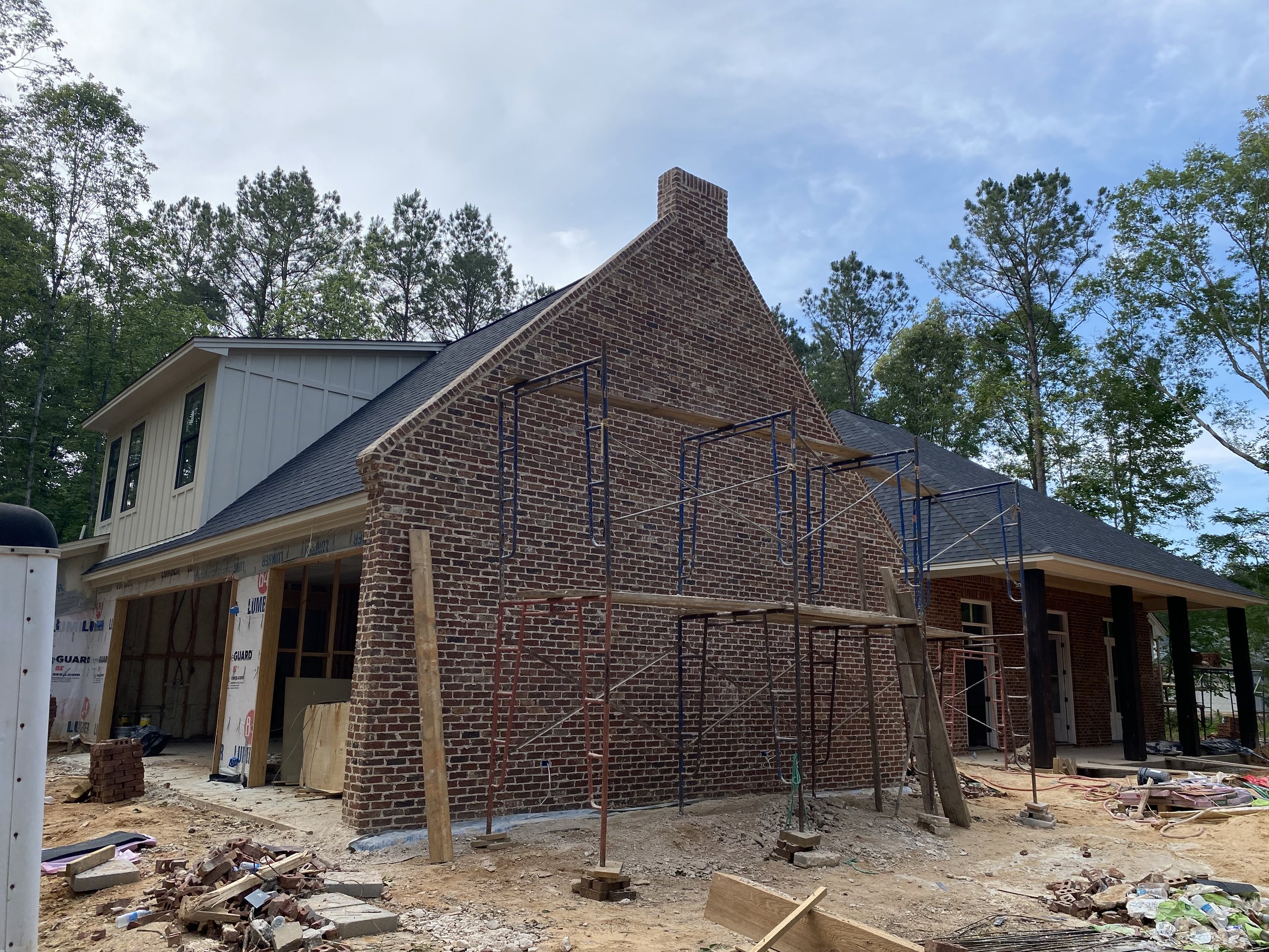 A house under construction, with brick walls, scaffolding, and construction materials on the ground, surrounded by trees under a partly cloudy sky.