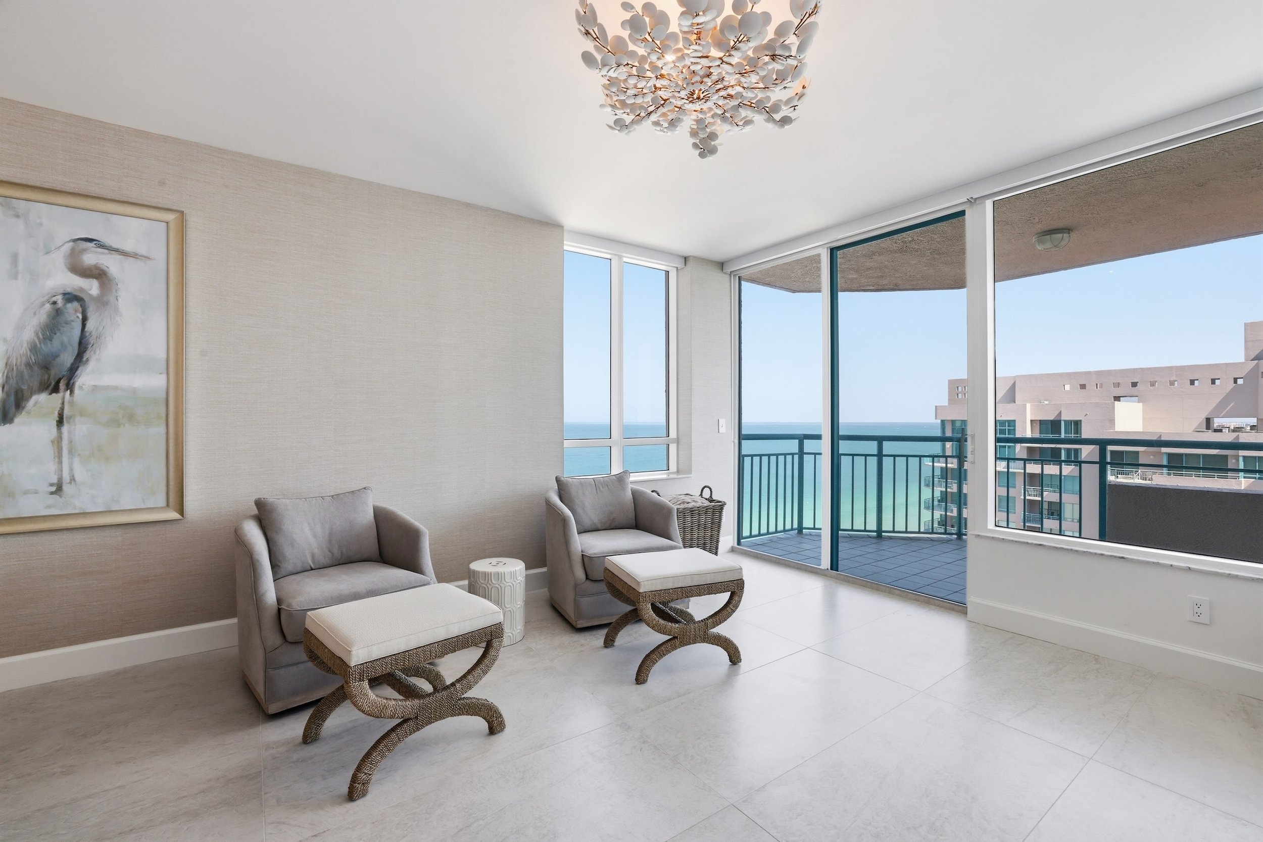 Living room with beige armchairs and ottomans, artwork of a heron on the wall, large windows with a view of a building and the ocean, and a ceiling light fixture.