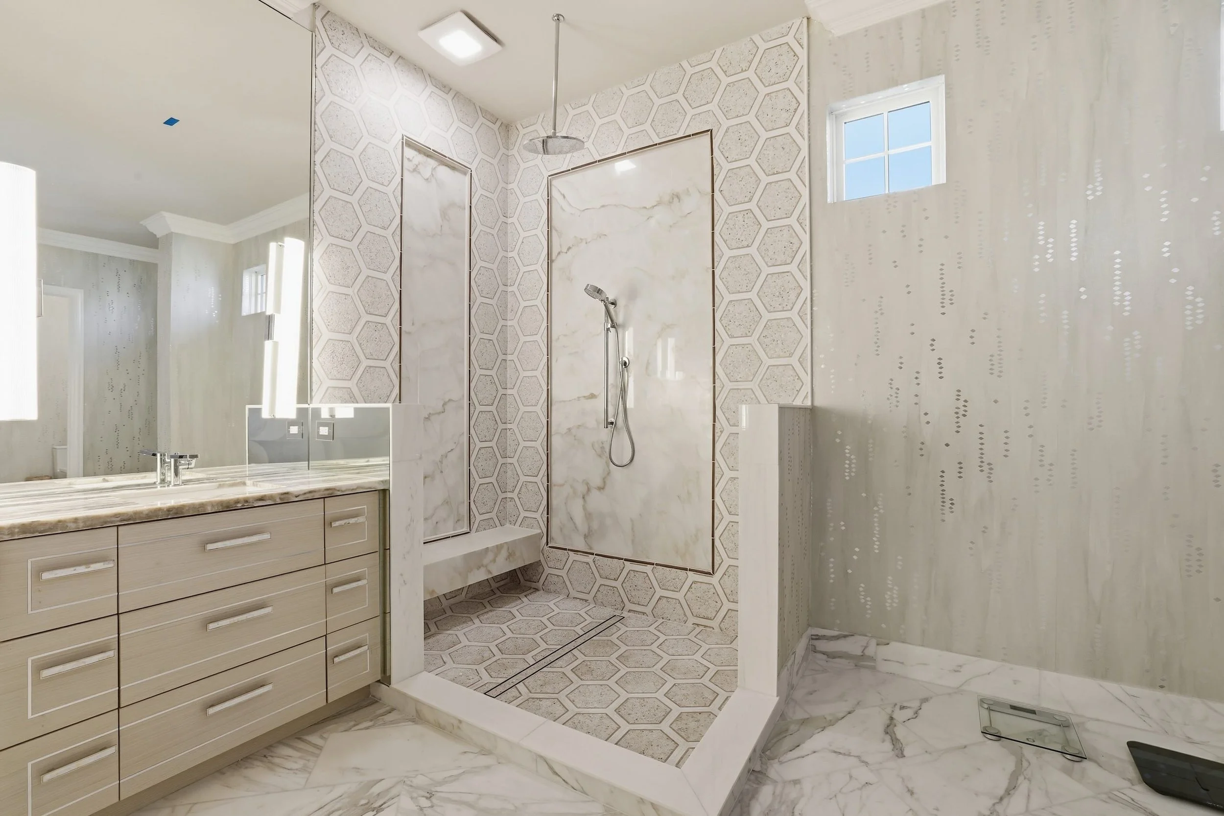 Modern bathroom with a walk-in shower featuring hexagonal and marble tiles, a beige vanity with drawers, a large mirror, and a window with blue sky visible outside.