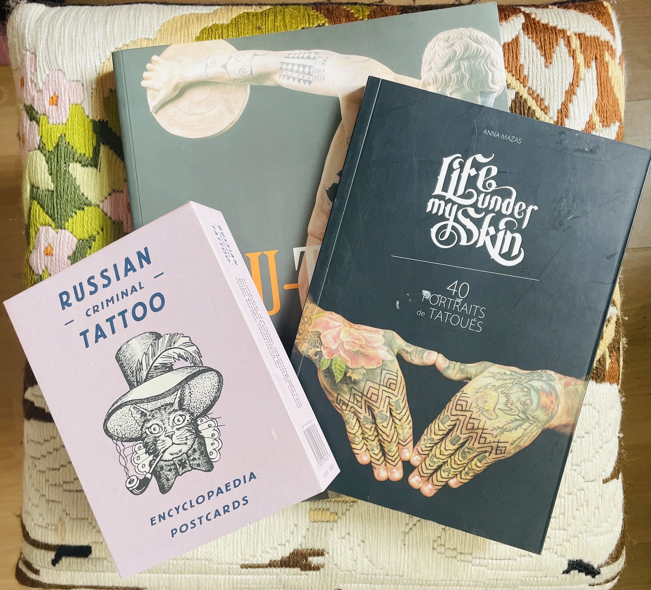 Several books, including one titled 'Life Under My Skin' by Anna Mazas, are resting on a patterned, multicolored fabric surface.