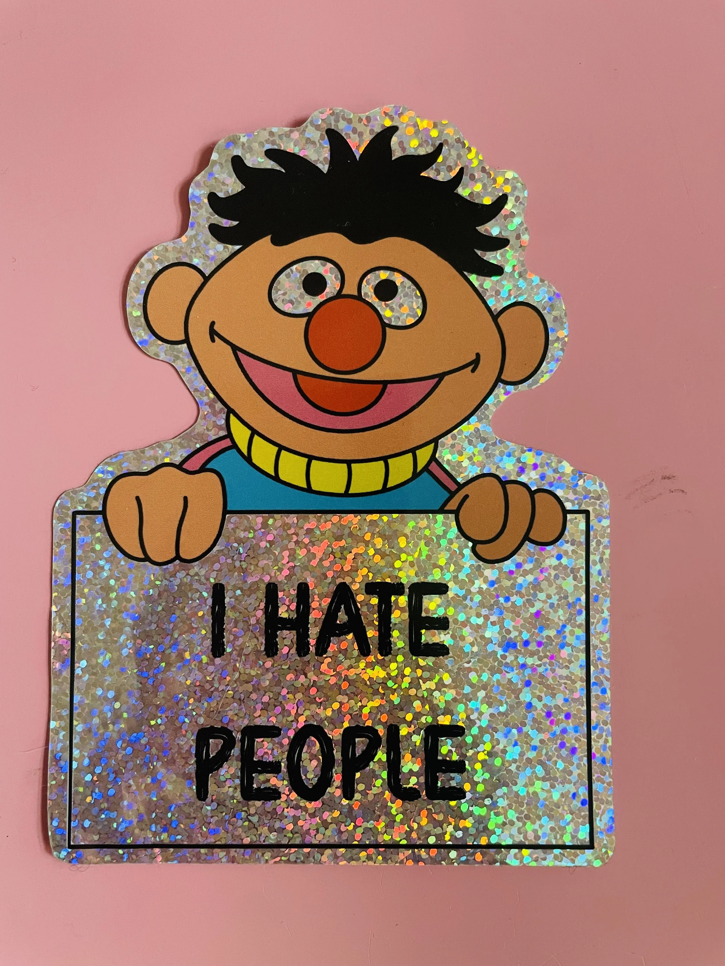 A sticker of a cartoon character with black hair, freckles, and orange nose, smiling, holding a sign that reads 'I HATE PEOPLE' in black letters, on holographic glitter background, on a pink surface.