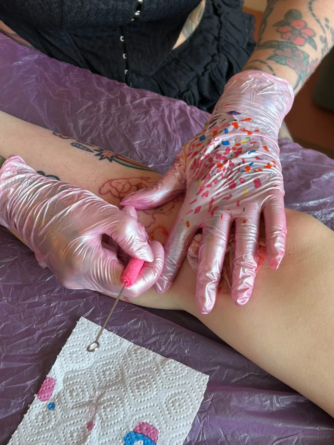 A tattooed person wearing pink gloves is applying ink to a customer's arm during a tattoo session. The person's arm has colorful tattoos, and the customer is lying on a purple cover with a paper towel nearby.