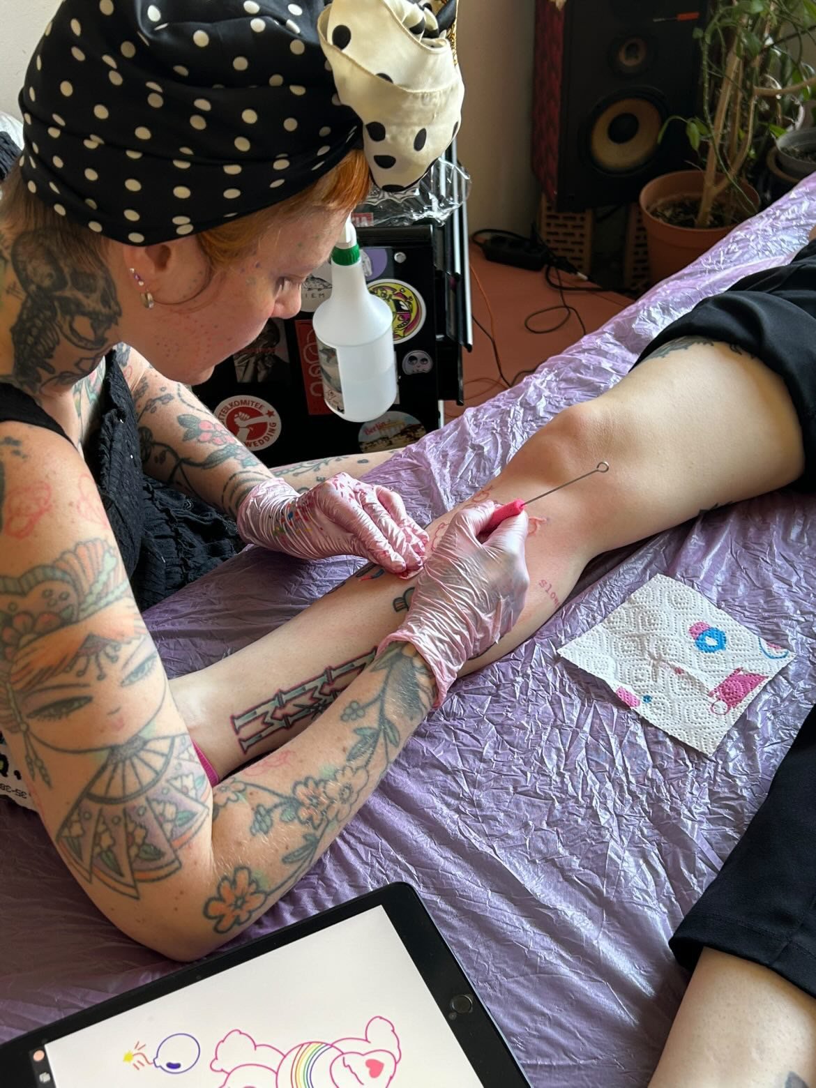 A tattooed woman administering a tattoo to a person's leg using a tattoo needle, with tattoo supplies and equipment nearby.