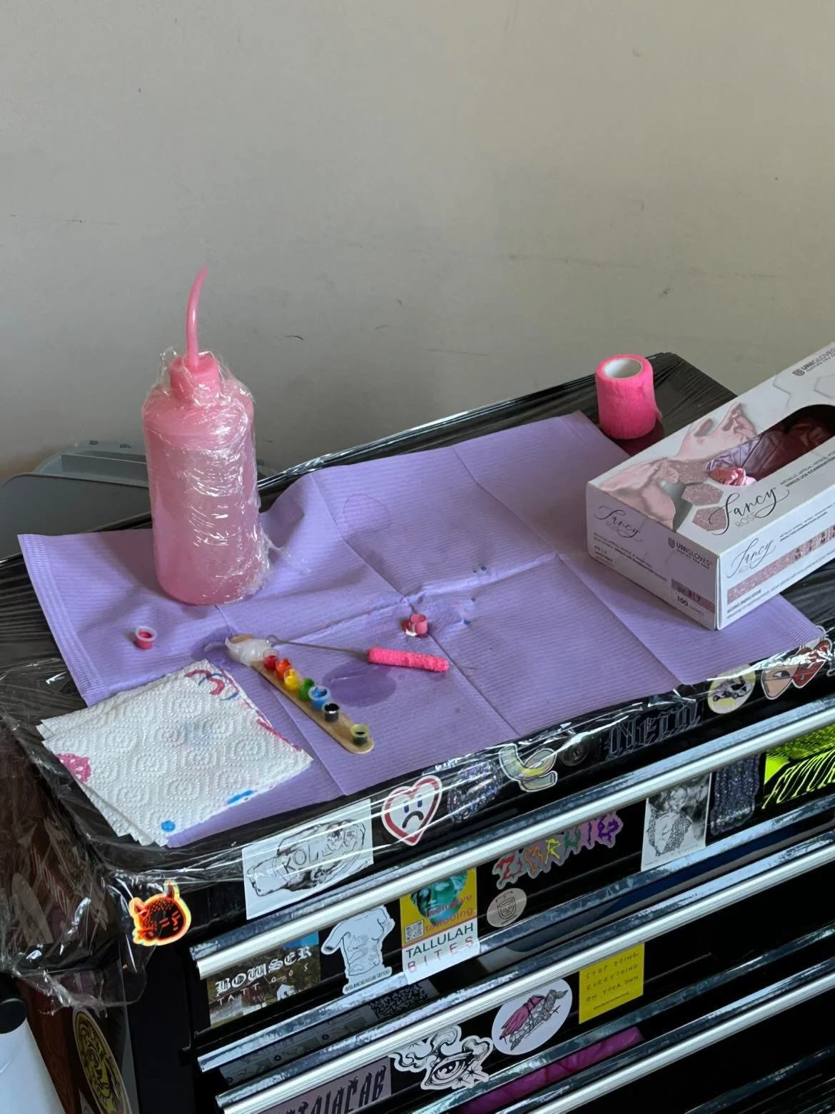 A small table with purple paper towels, a pink candle, skincare or makeup supplies, a box of tissues, and stickers on the front, including a white paper towel and a small palette of colorful nail polish.