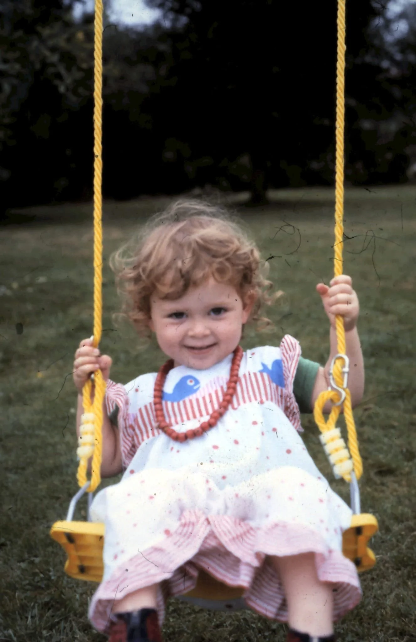 Young child with red curly hair smiling while sitting on a yellow outdoor swing in a park during daytime.
