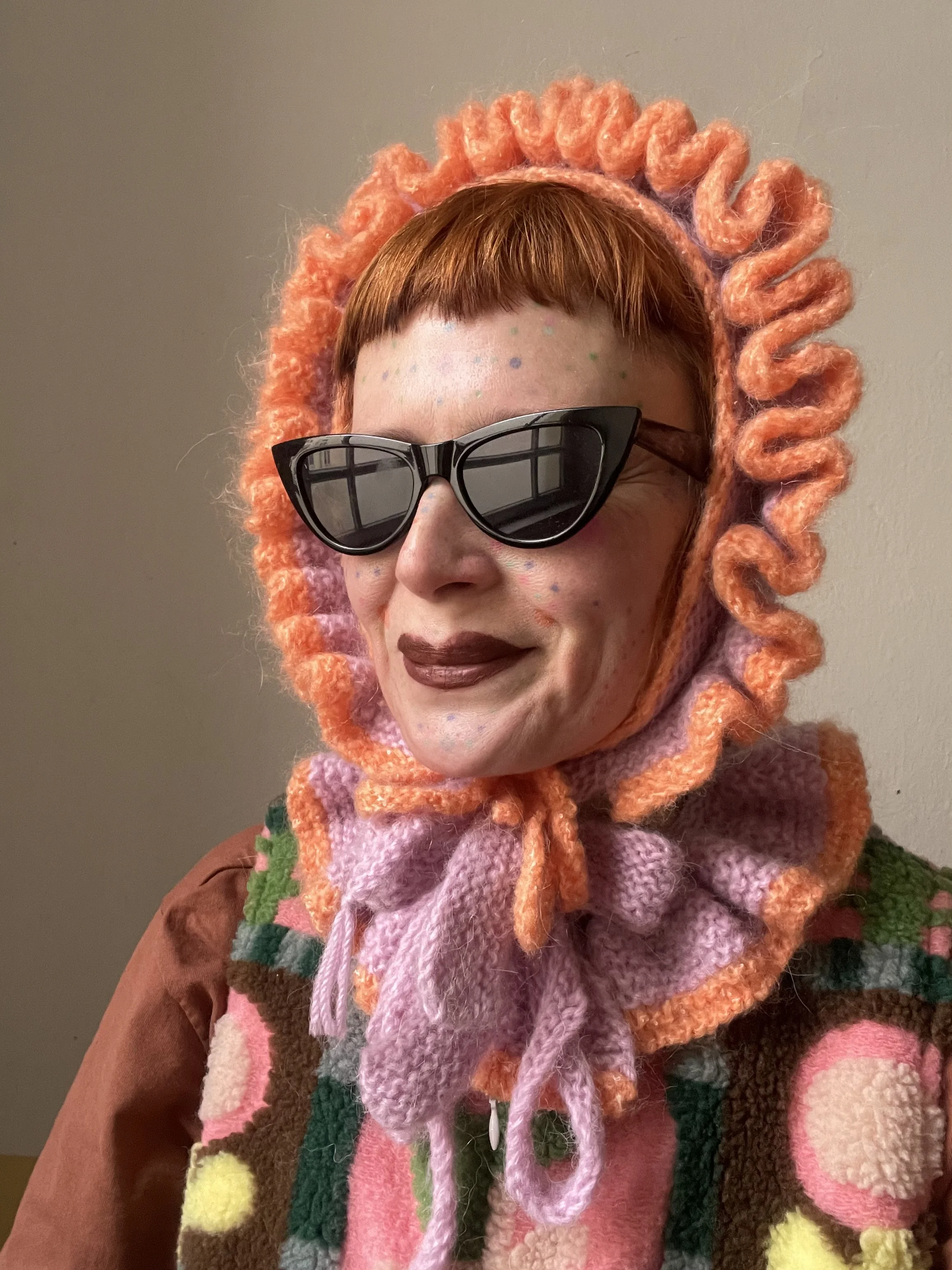 Person wearing black cat-eye sunglasses, a colorful handknitted bonnet, and a multicolored patchwork sweater, standing against a plain background.