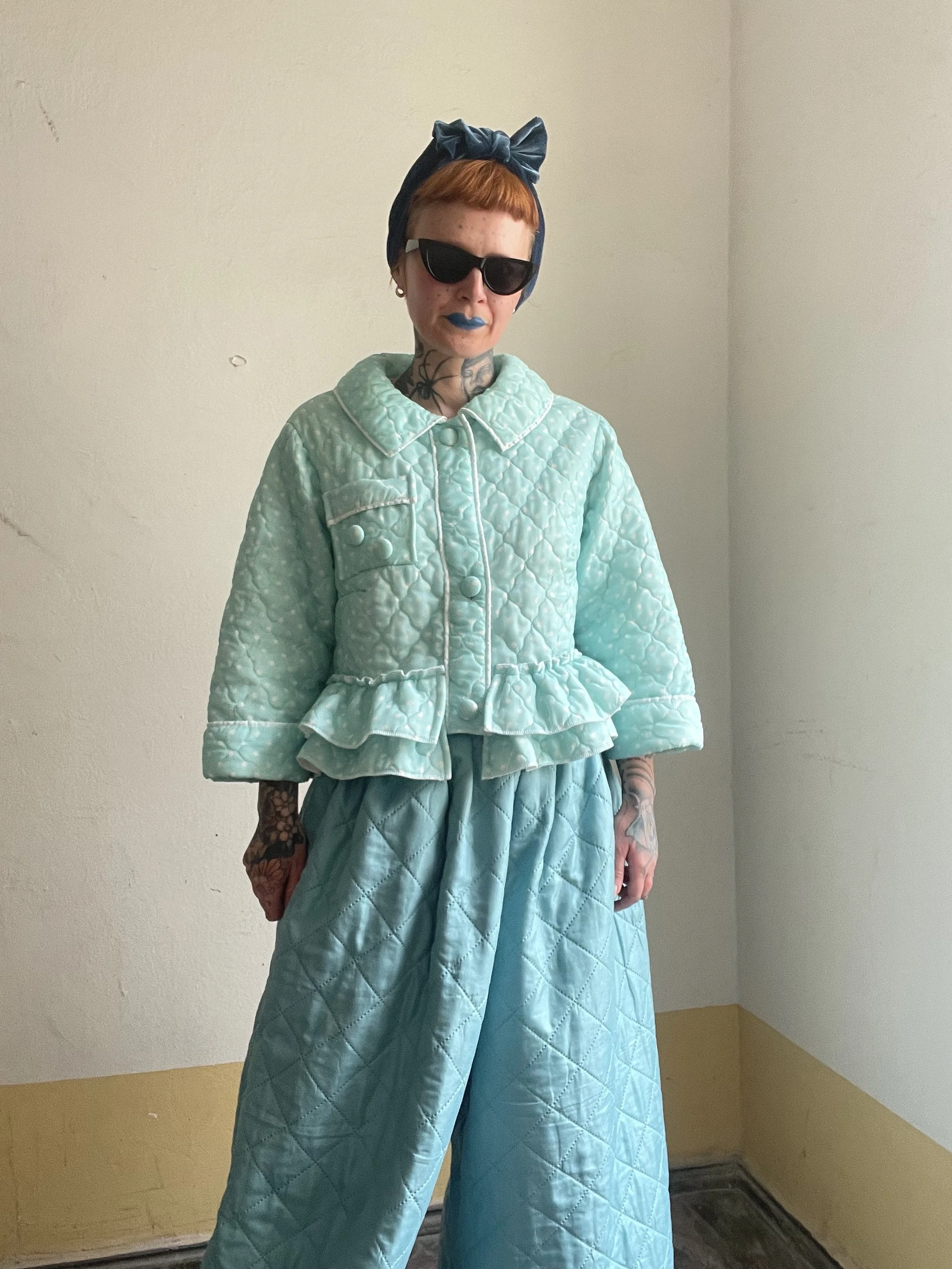Woman with tattoos wearing black sunglasses, a blue headscarf tied in a bow, a mint green quilted jacket, and matching wide-leg pants, standing in a corner of a room with beige and white walls.