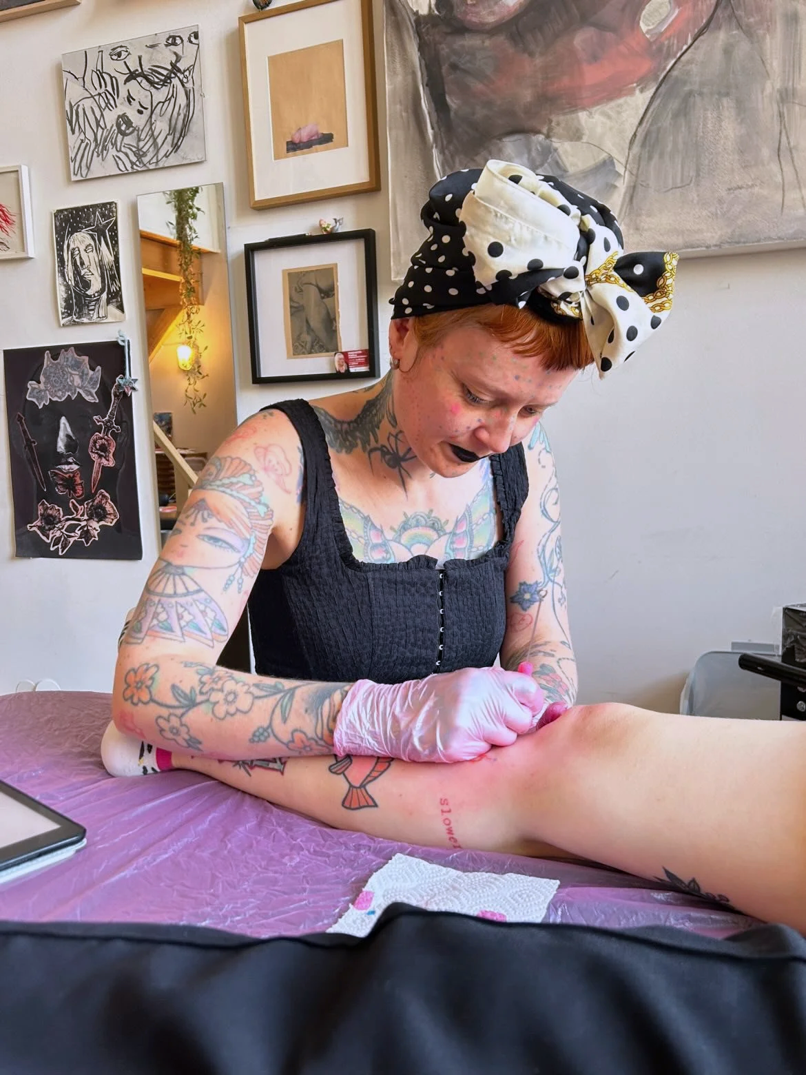 A tattooed woman with short red hair and a polka dot headscarf tattooing a person's leg in an art gallery or tattoo studio.