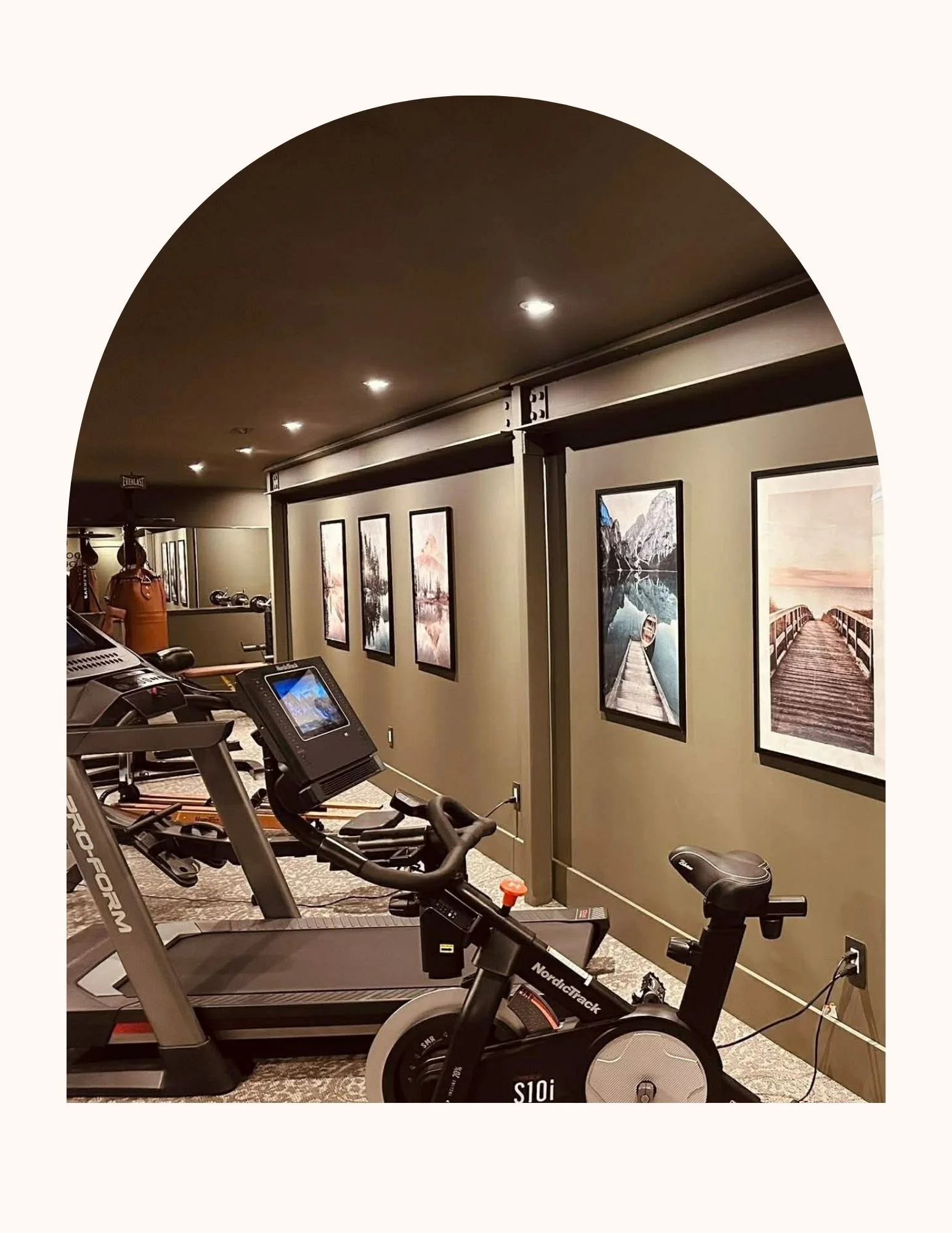 The image shows a fitness room with two exercise bikes in the foreground and a treadmill. The wall is decorated with framed landscape photographs and there is a coat rack with jackets and umbrellas in the background.