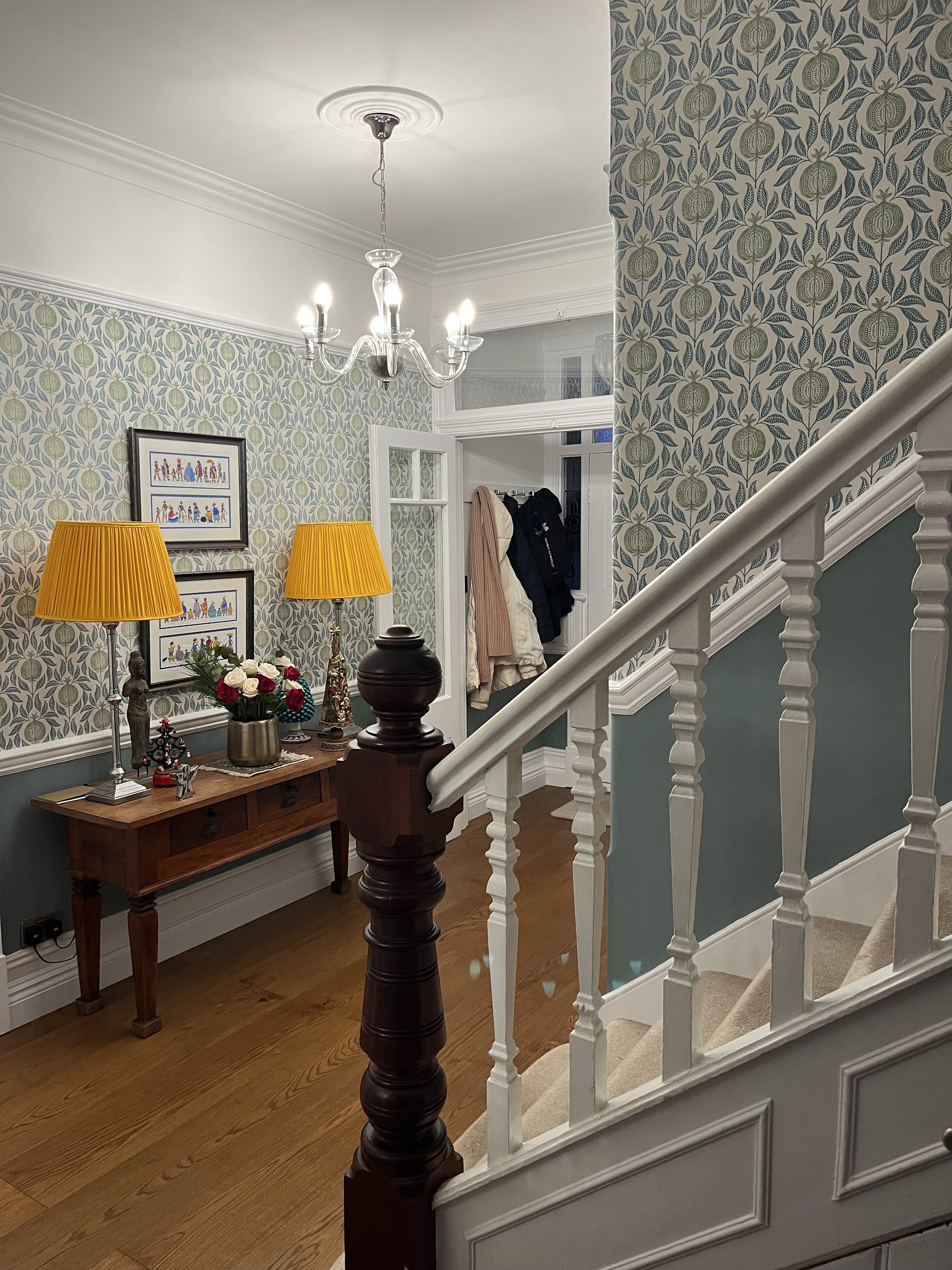 View of a cozy foyer with patterned wallpaper, a small wooden table with a bouquet of red and white roses, two yellow lamps, framed artwork, a chandelier, and a staircase with white spindles and a beige carpeted stairs leading upstairs. Coats and a bag are hanging near a doorway.