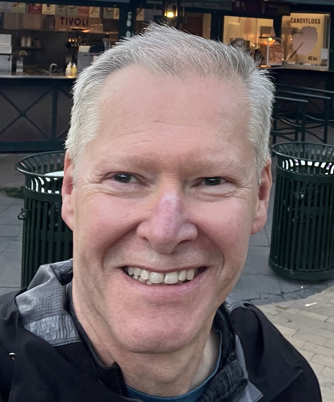 Close-up of a smiling man with short gray hair, showing his teeth, standing outdoors at night near a restaurant or bar with black trash cans and signs in the background.