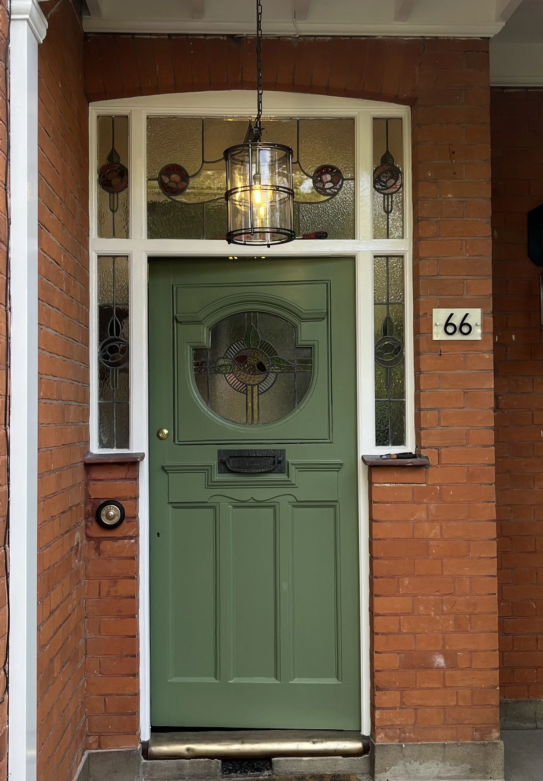 Green front door with a stained glass window, surrounded by brick walls and glass panels, outdoor light fixture with an exposed bulb, doorbell, and mail slot.