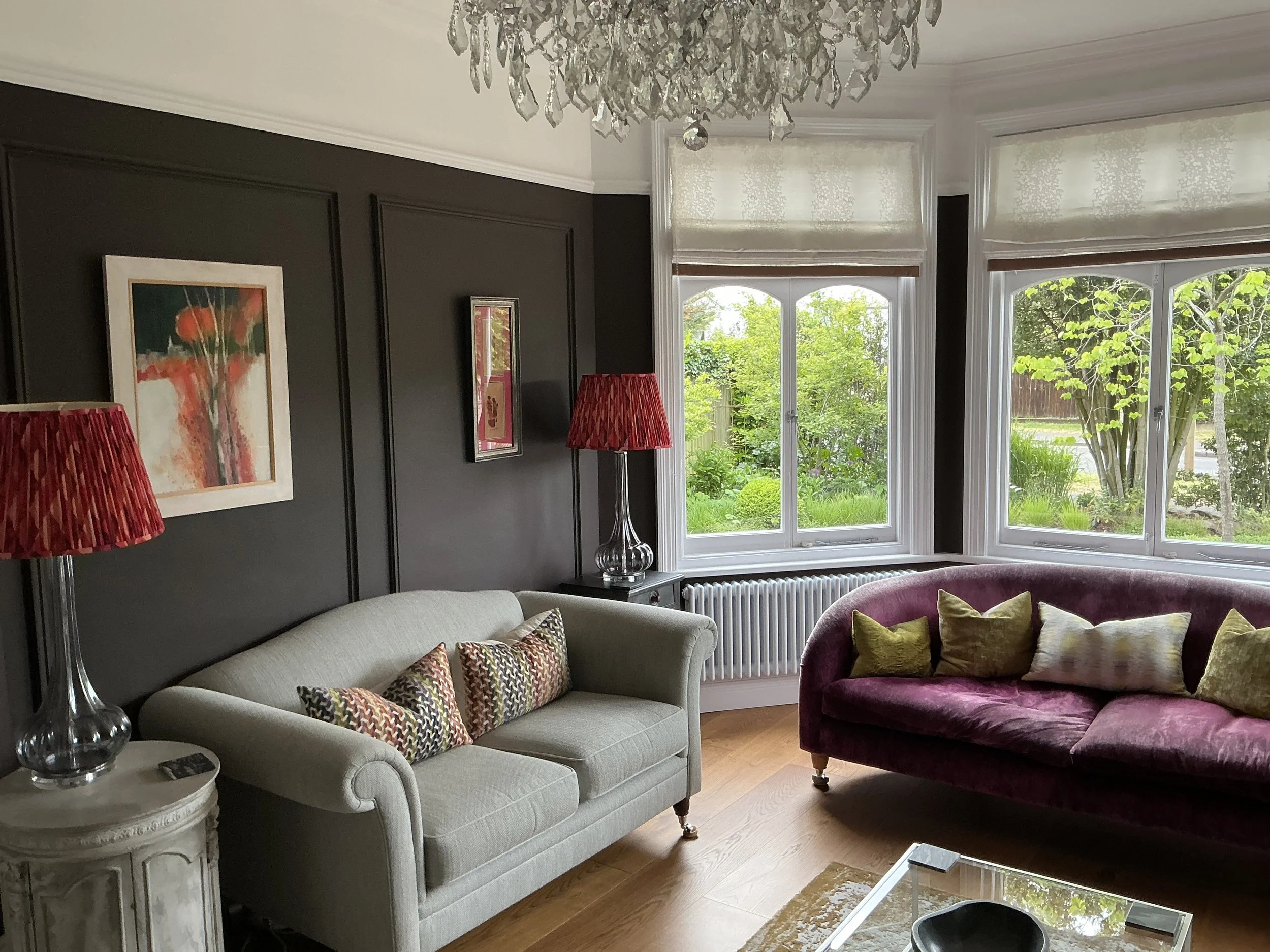 A living room with large bay windows showing green trees outside. The room has dark-colored walls with artwork and two red lamps. There is a beige sofa with patterned pillows, a burgundy velvet armchair, a white side table, and a glass-top coffee table.