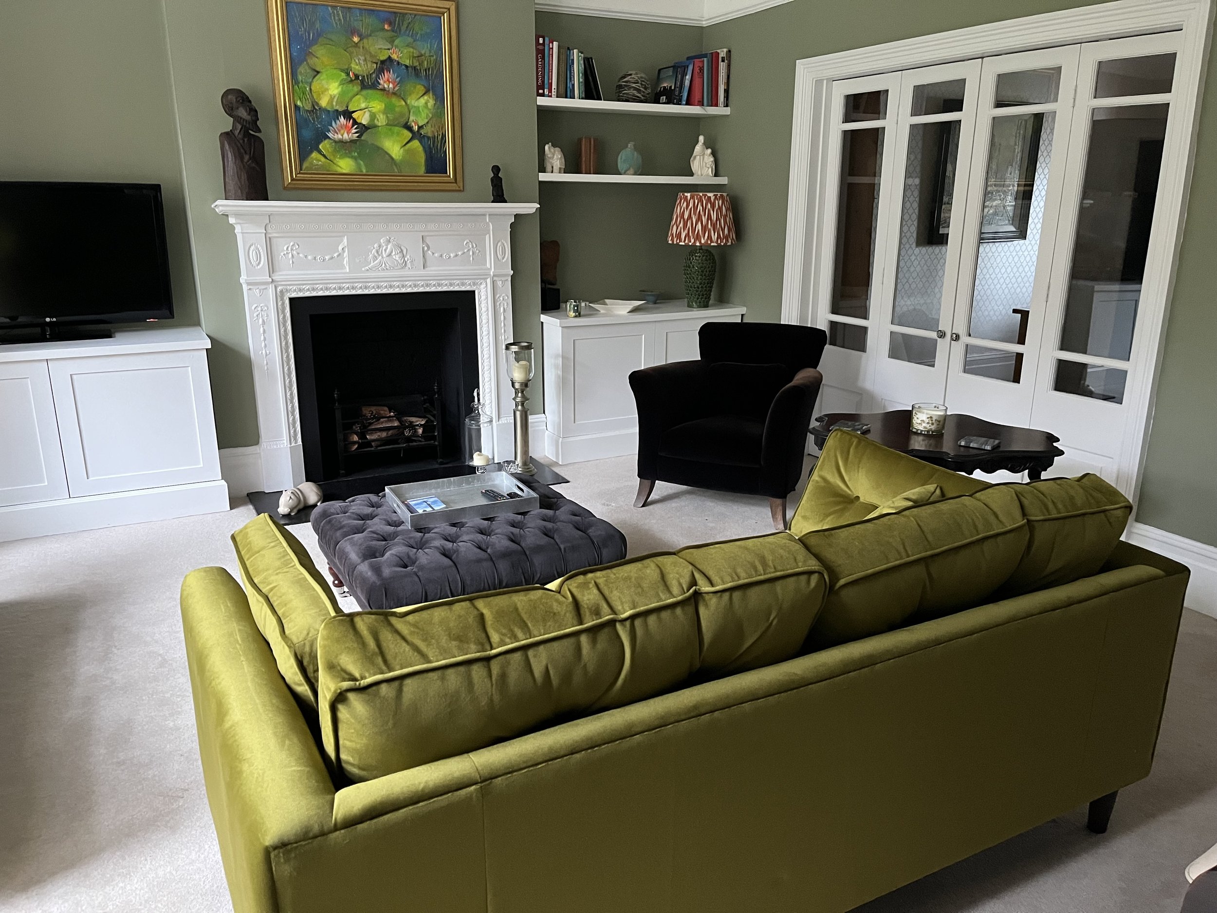 Living room with green velvet couch, black armchair, white fireplace with decorative carvings, and artwork of water lilies above the fireplace. There are two white shelves with books and decorative items. A large window with white divided panes is on the right side of the room.