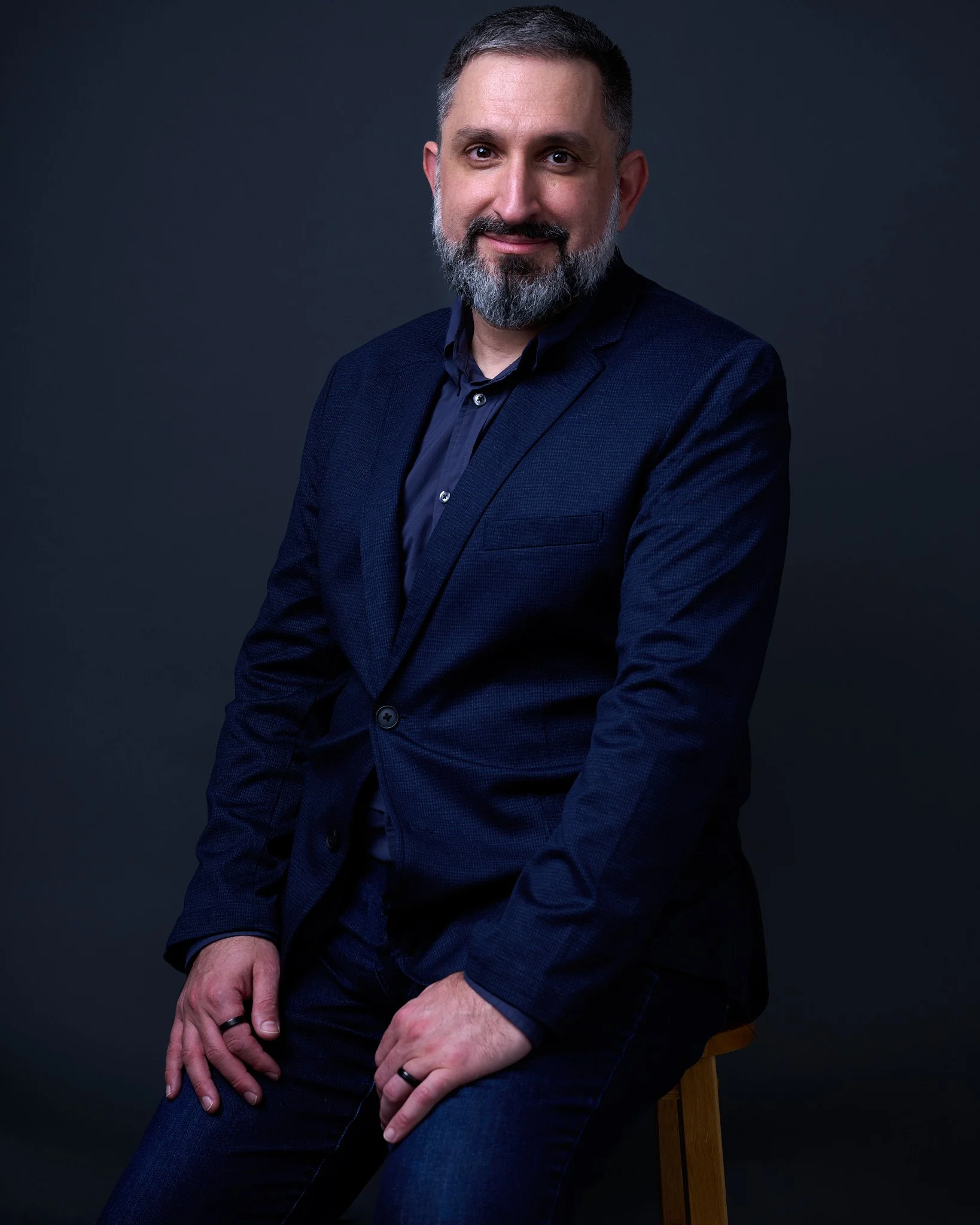 A man with gray hair and a beard sitting on a chair against a dark background, wearing a dark blazer and shirt, with his hands resting on his lap.