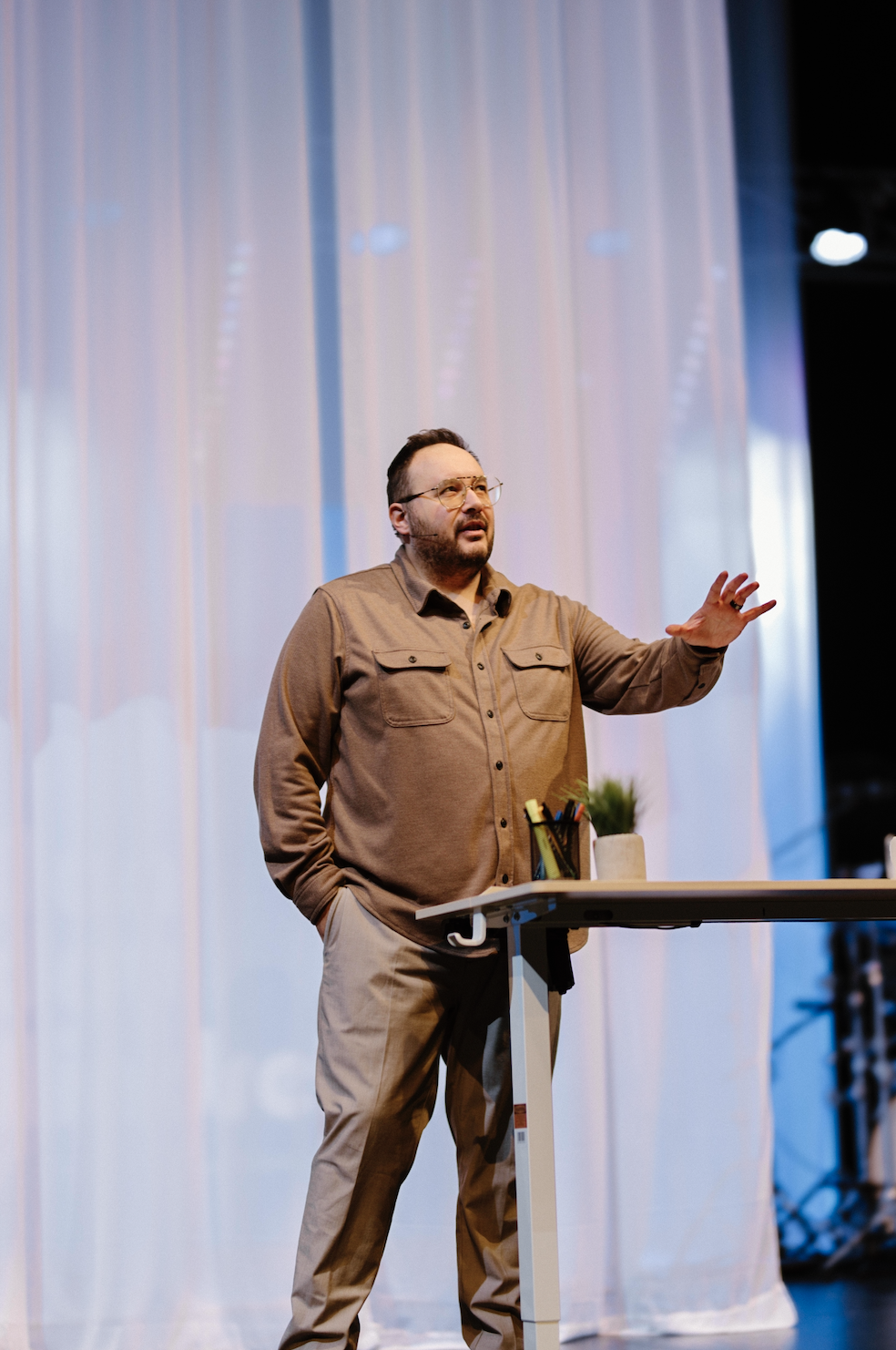 A man in glasses and a tan shirt stands behind a white table, speaking with one hand raised, in front of a large, blurred curtain backdrop and stage lighting.