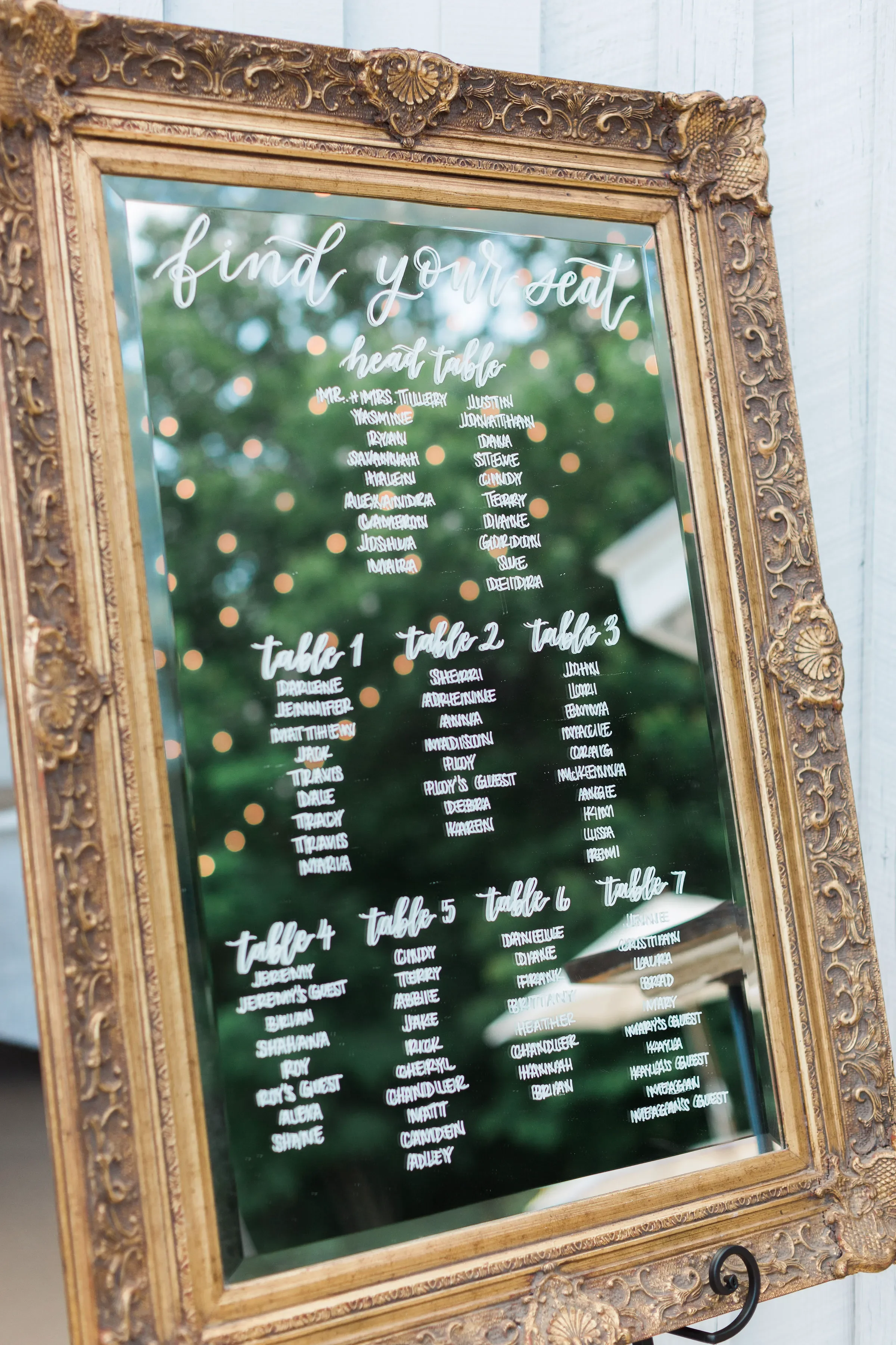 Guide your guests beautifully with a custom mirror wedding seating chart featuring elegant hand-lettered white calligraphy. This large vintage gold-framed mirror sign reads "find your seat" and lists a head table alongside tables one through seven. S