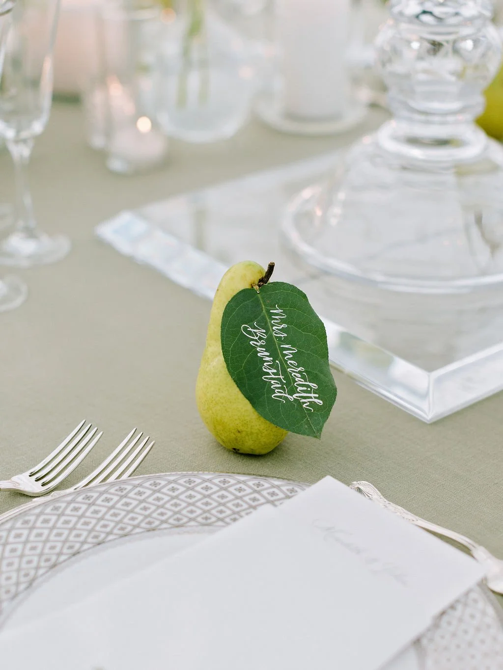 Hand-lettered calligraphy place card on a pear leaf attached to a fresh pear at an elegant wedding reception table.