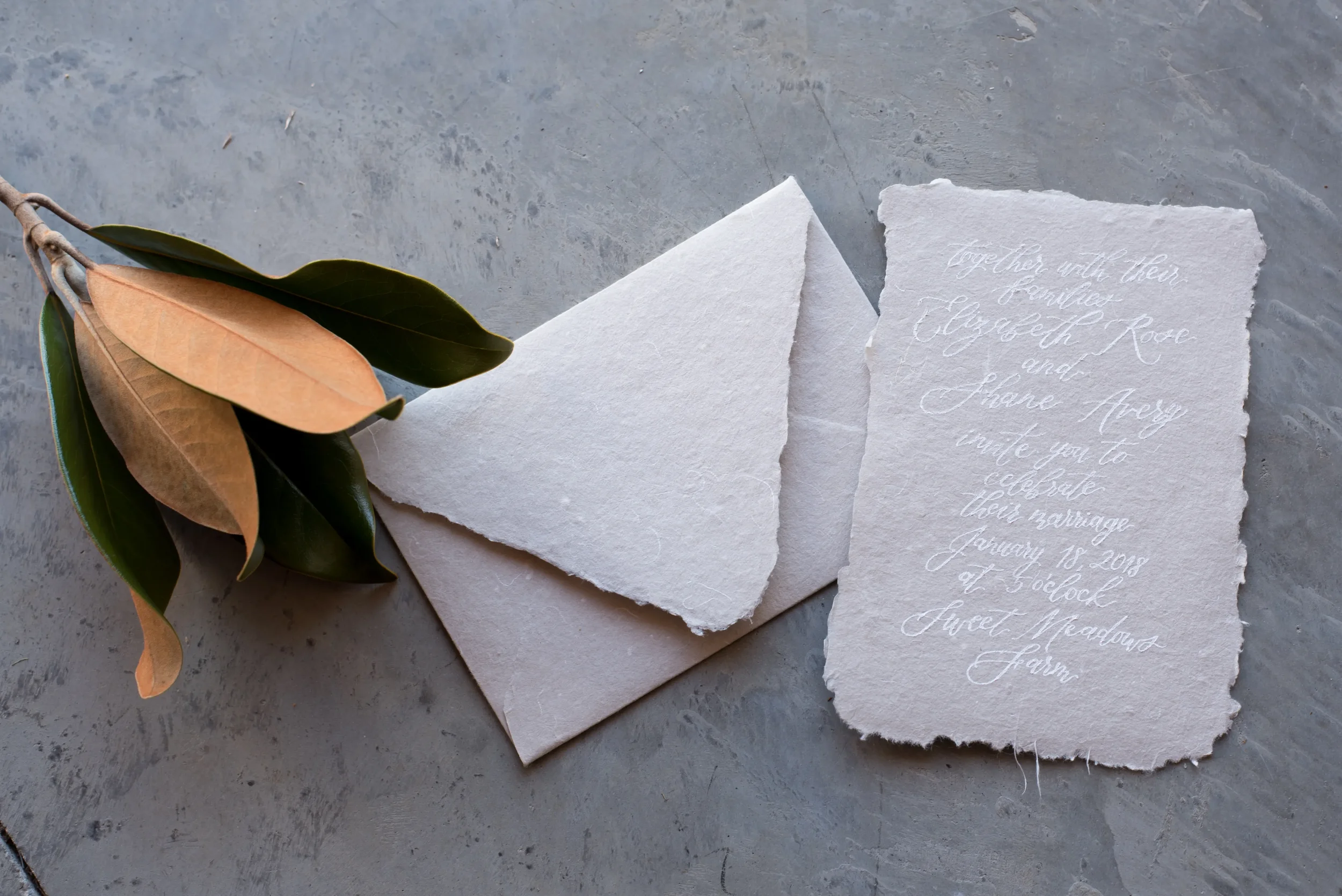 Deckled-edge handmade paper wedding invitation written in white script next to a euro flap envelope and fresh magnolia leaves.